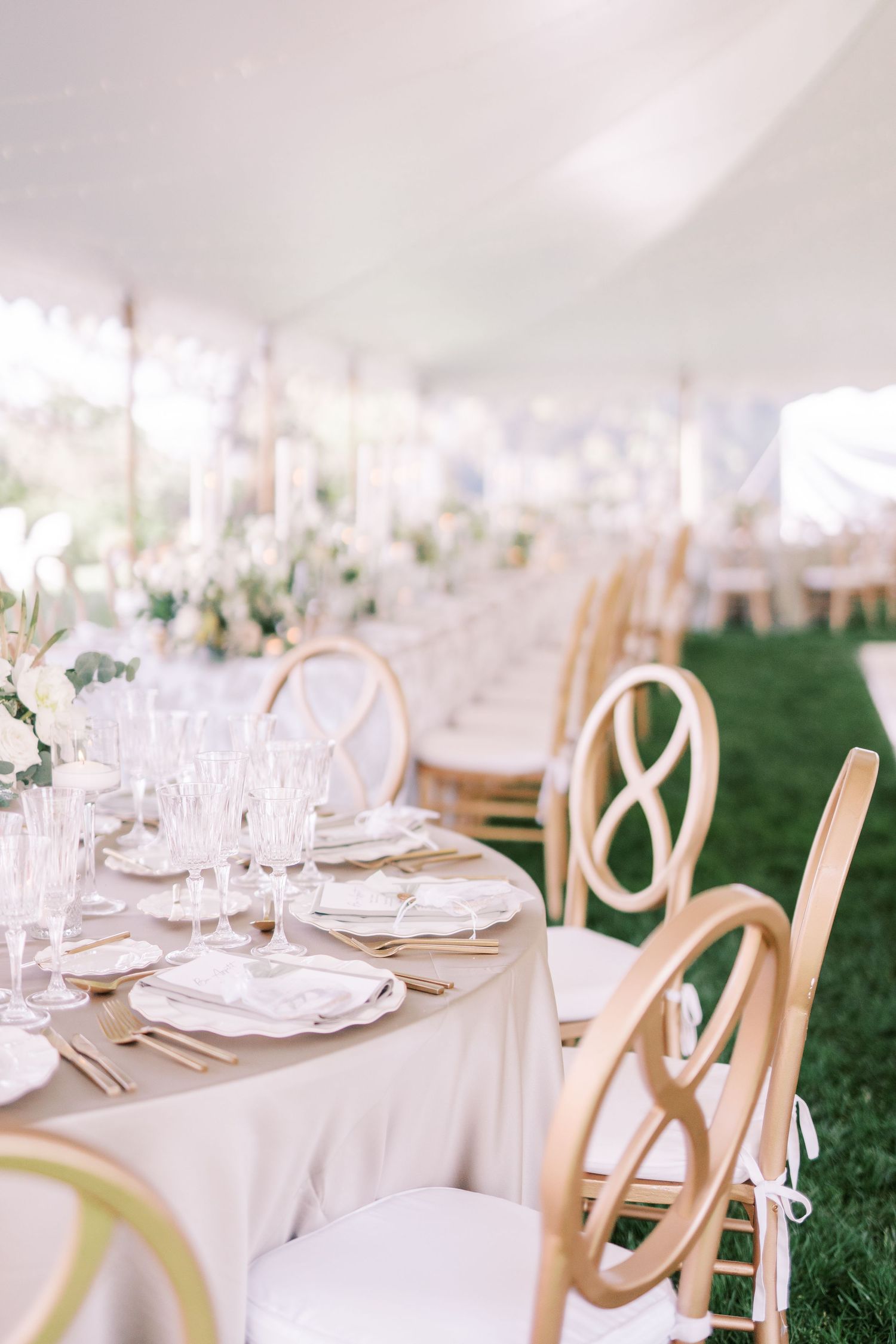 Elegant wedding reception table setting with light linens, crystal glassware and wooden crossback chairs on grass.