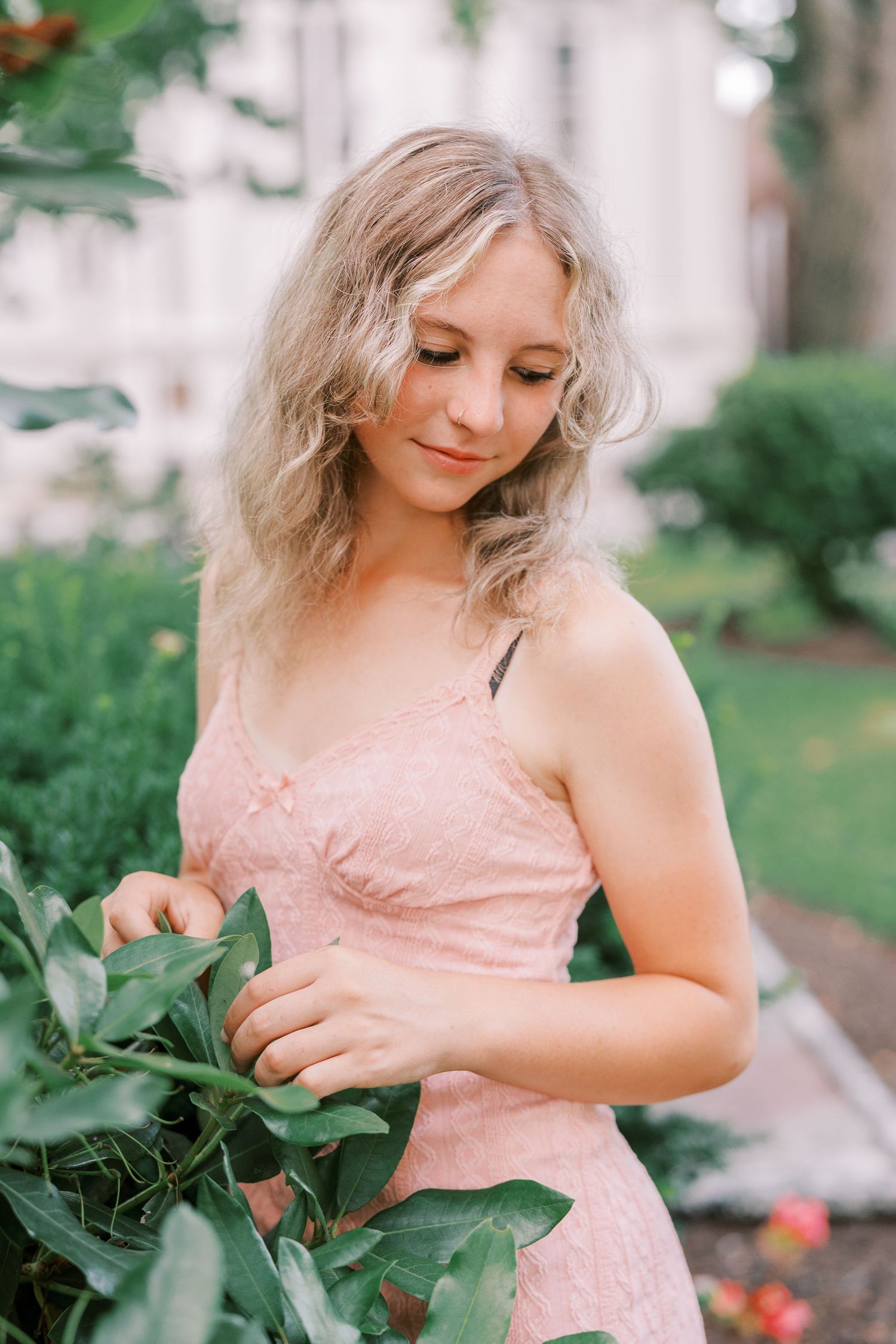 Dreamy outdoor portrait of woman in peach-colored dress gently touching leaves in a garden setting.