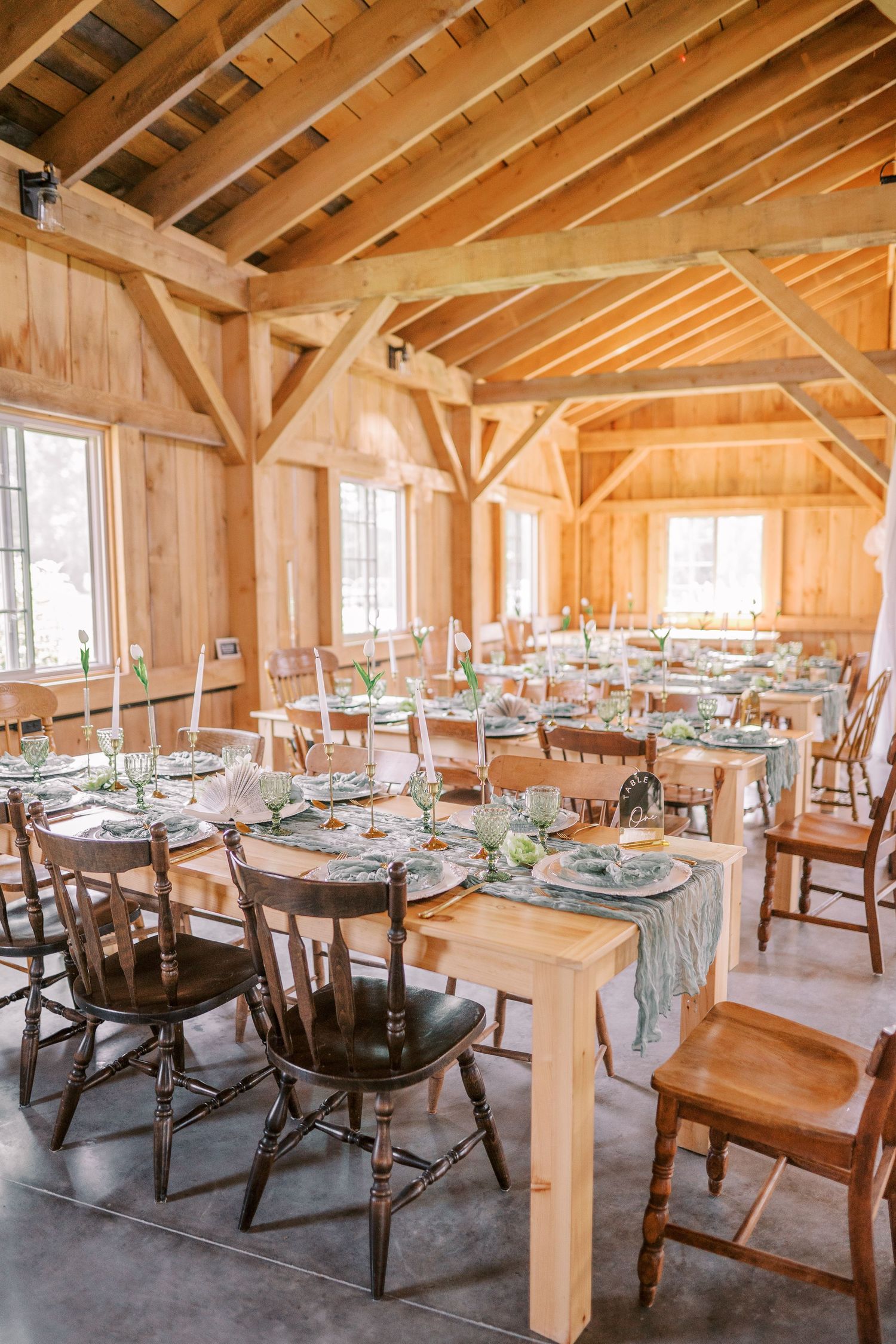 Light-filled rustic barn venue with wooden beams and tables set for a wedding reception with natural decor.