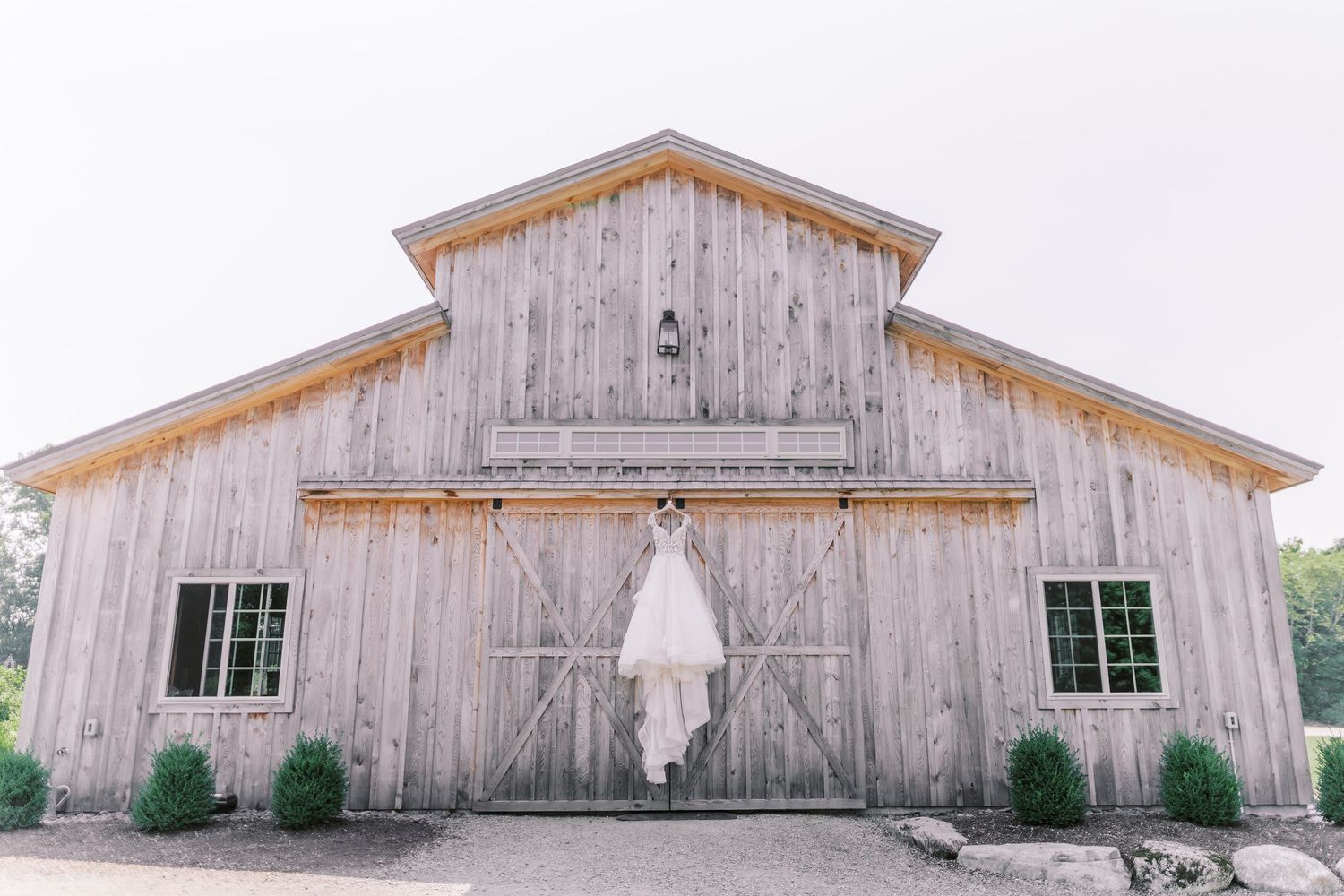 A rustic wooden barn with weathered gray siding features a white wedding dress hanging on its front doors.