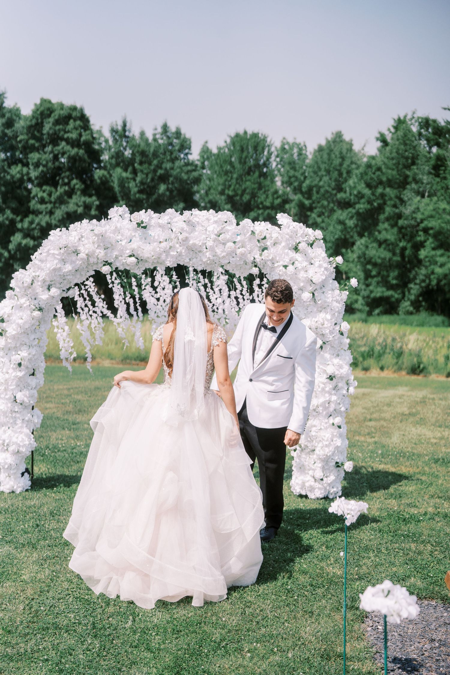 A beautiful outdoor wedding ceremony setup with an arch made of white floral arrangements leading down a path in a garden setting.