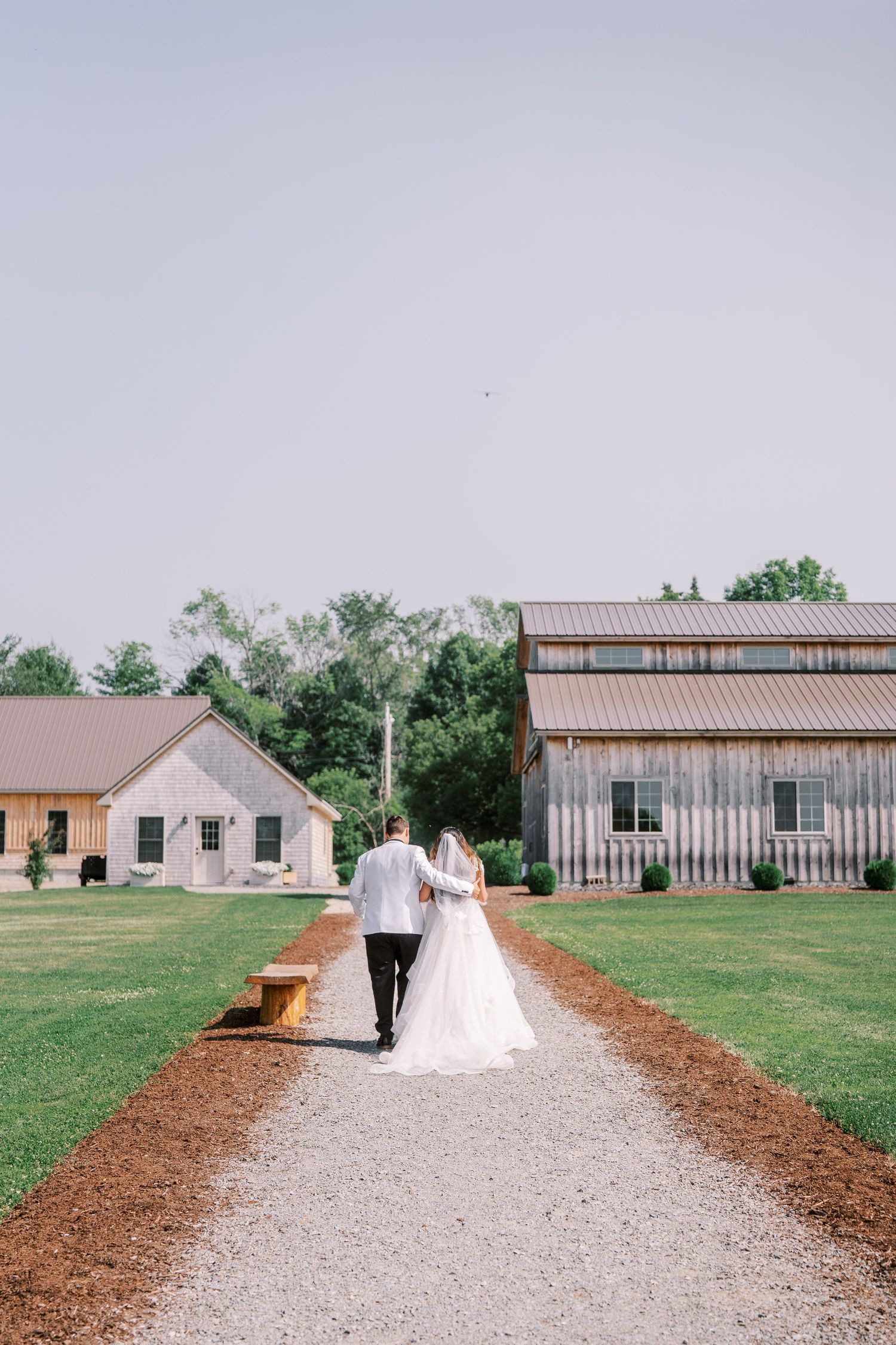Couple walking down gravel path between rustic barn buildings at countryside wedding venue on cloudy day.