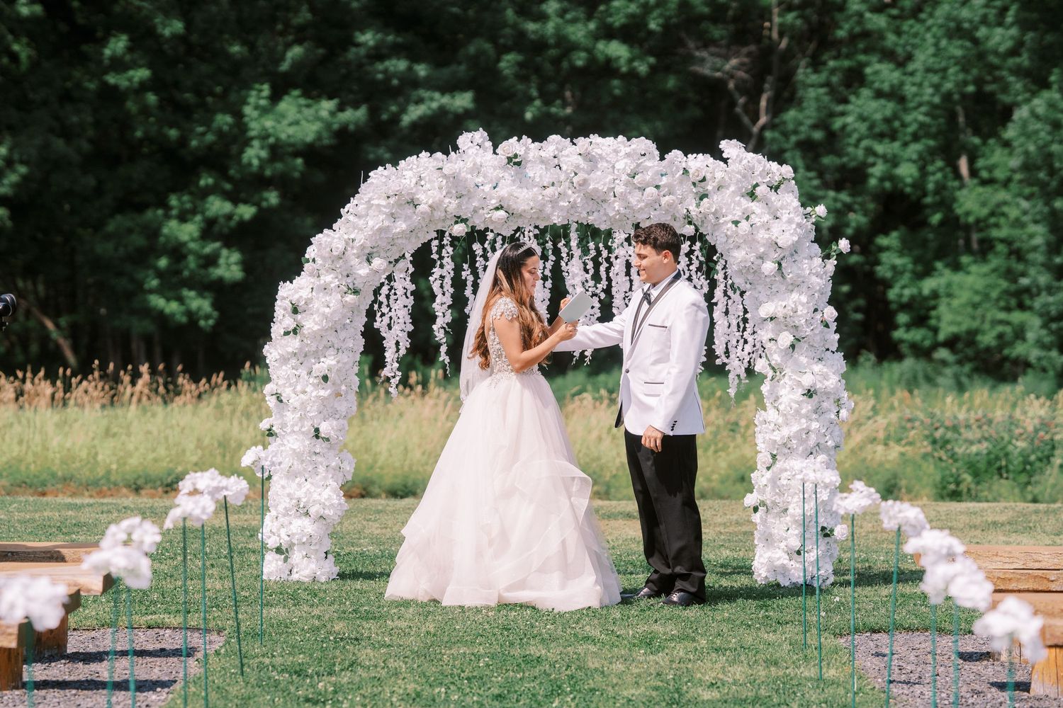 A beautiful outdoor wedding ceremony takes place under a stunning white floral arch surrounded by lush greenery.
