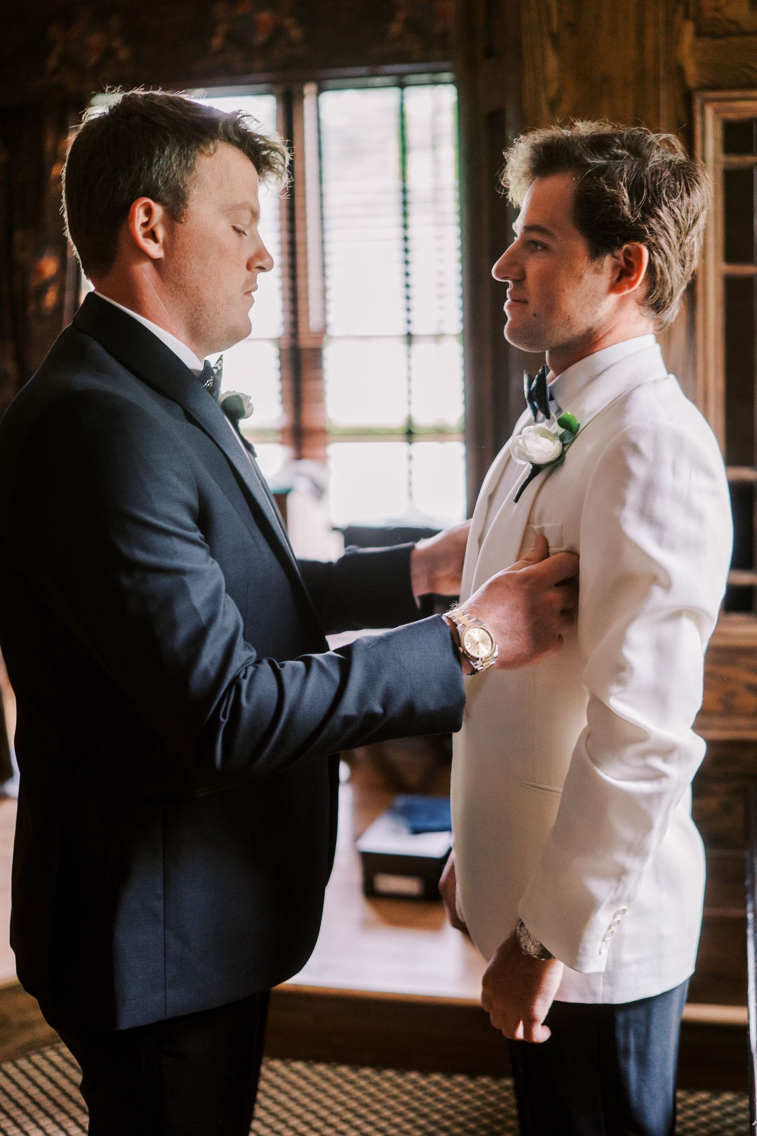 Two people in formal attire help each other prepare for a special occasion in a warmly lit rustic room.