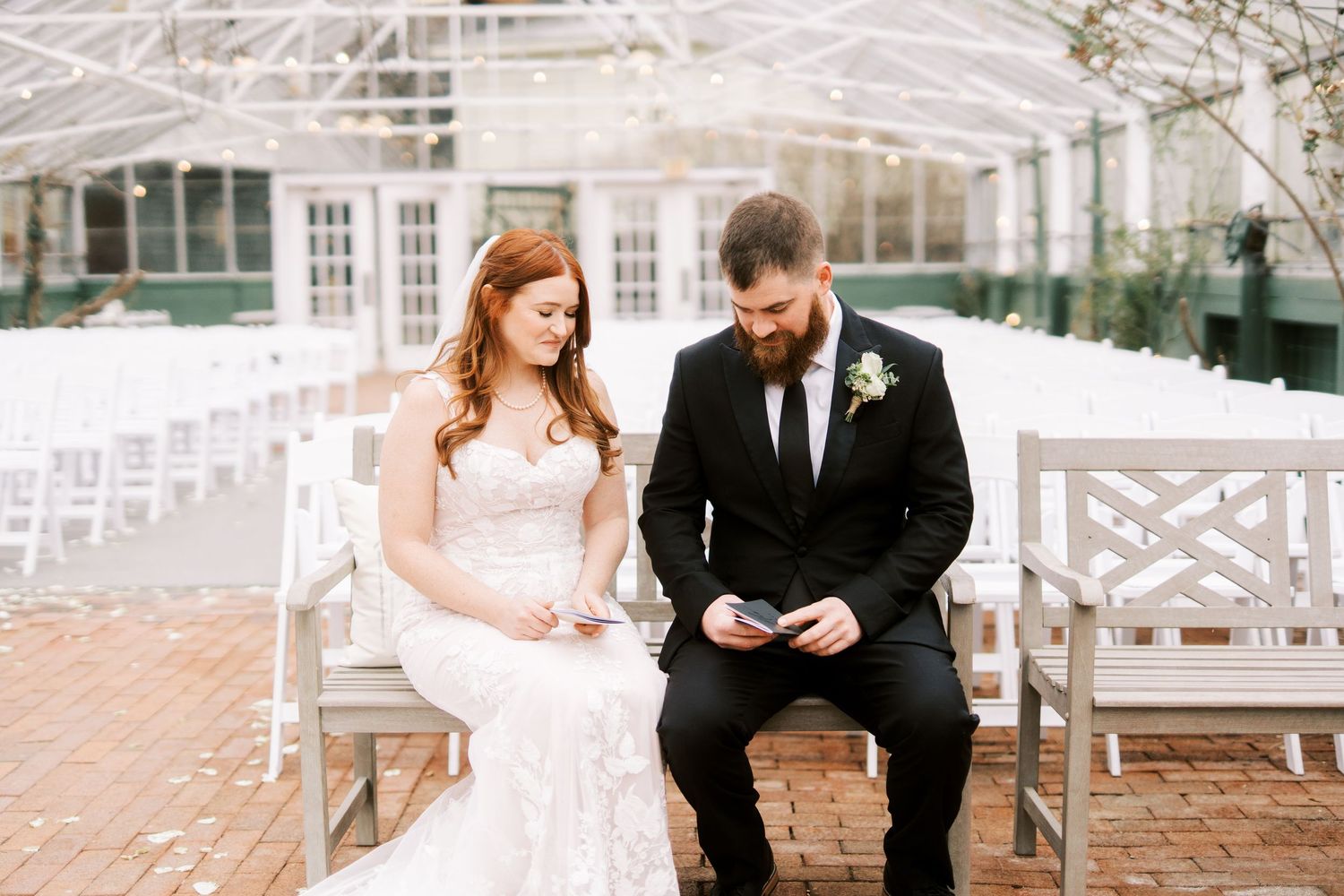 Couple sharing an intimate moment on a wooden bench inside a greenhouse venue with white chairs.