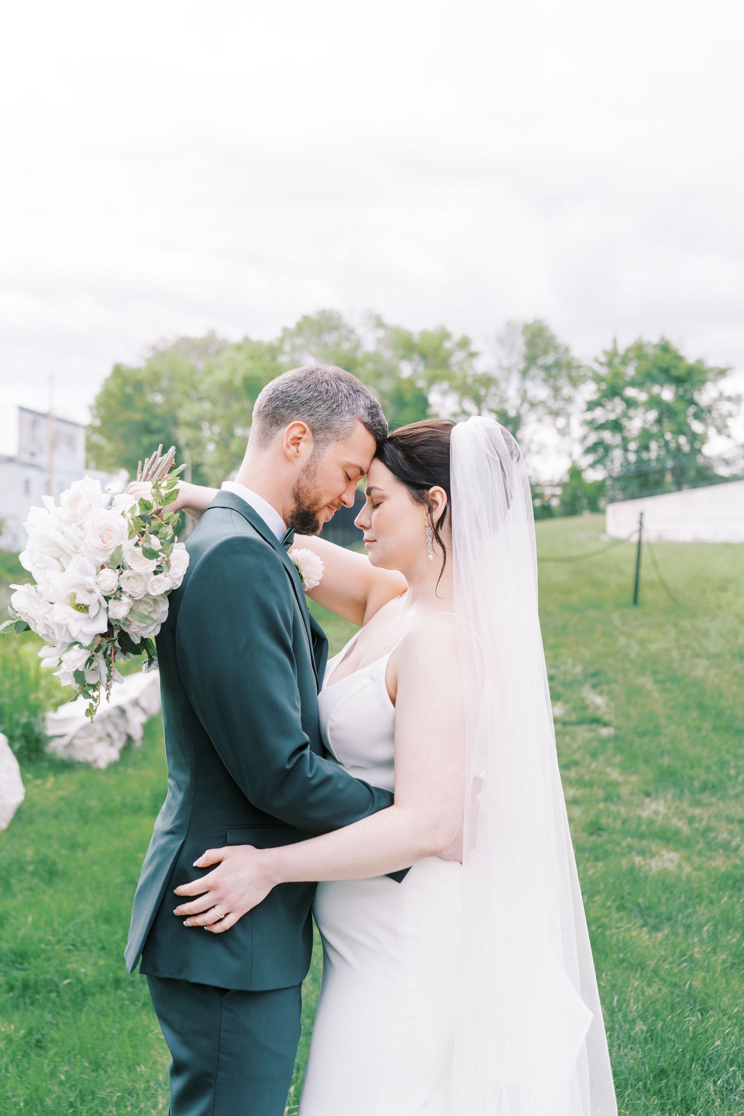 Newlyweds share an intimate moment outdoors with white floral bouquet and flowing veil.