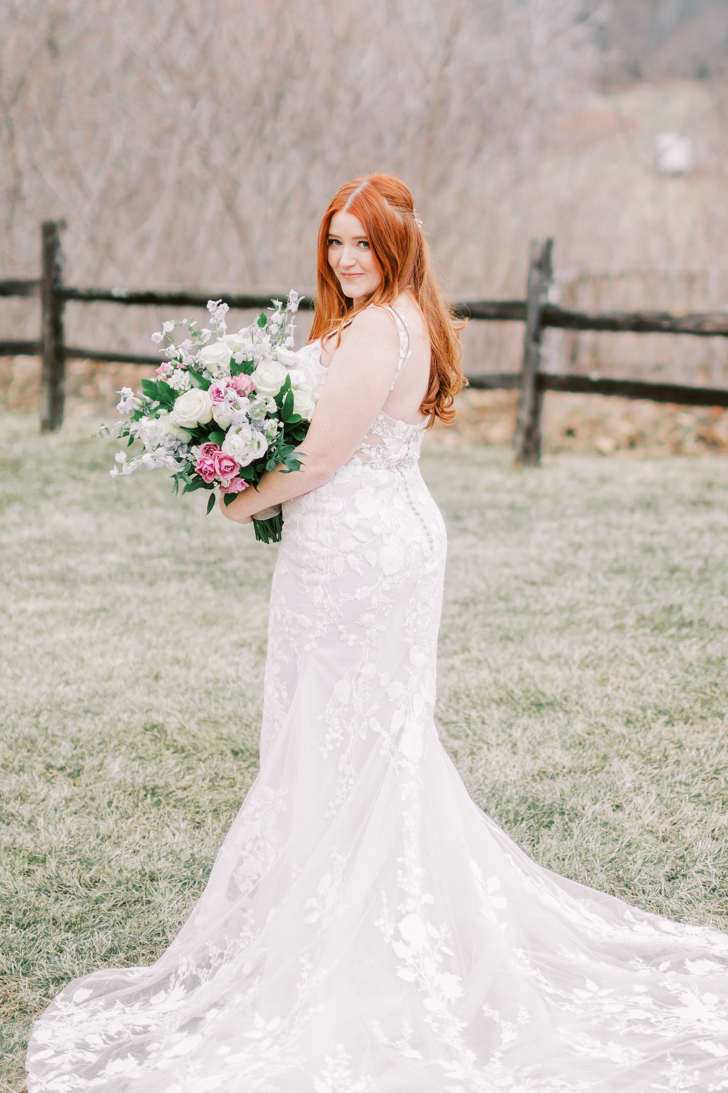 Bride in lace wedding gown holding floral bouquet poses in front of rustic fence.