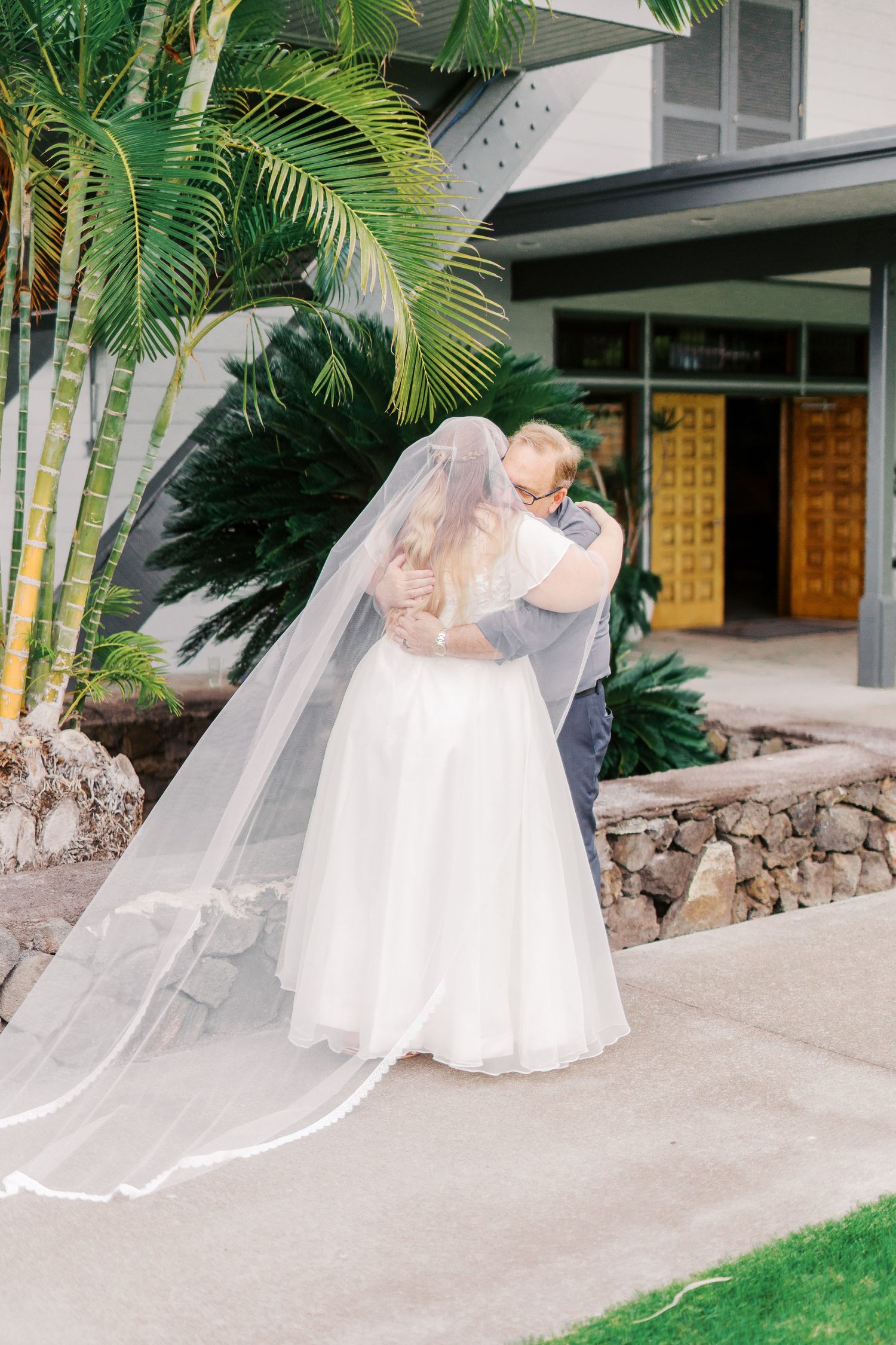 A romantic wedding moment captured outside a tropical venue with palm trees and stone walls.