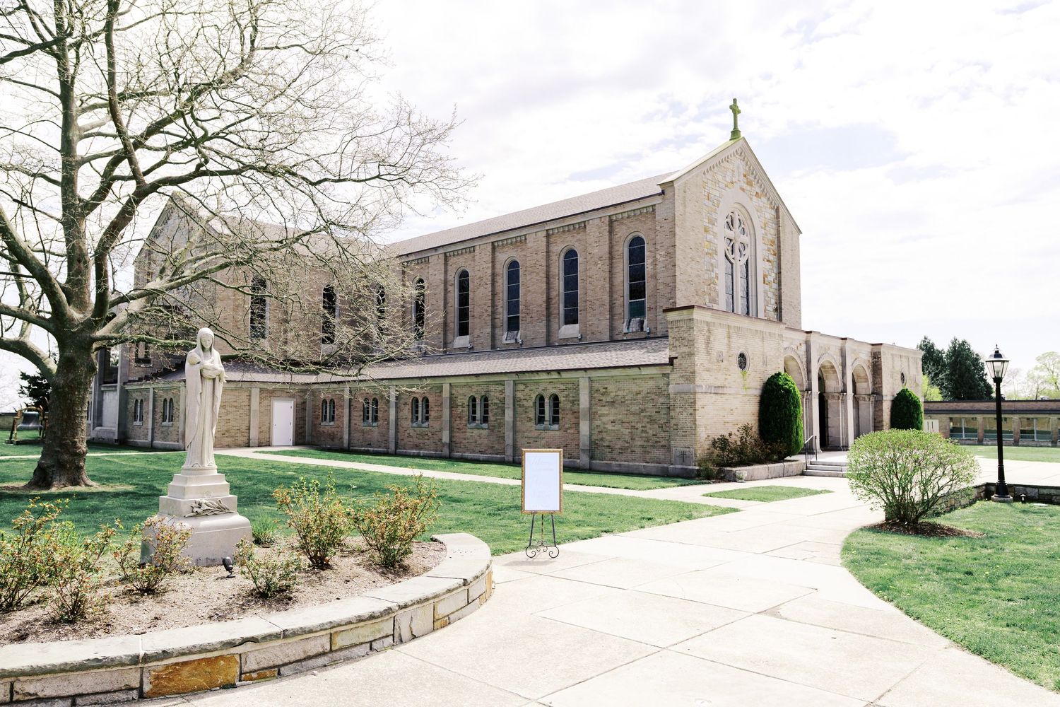 A stone Catholic church building with a cross on top stands next to a curved sidewalk and landscaped grounds.