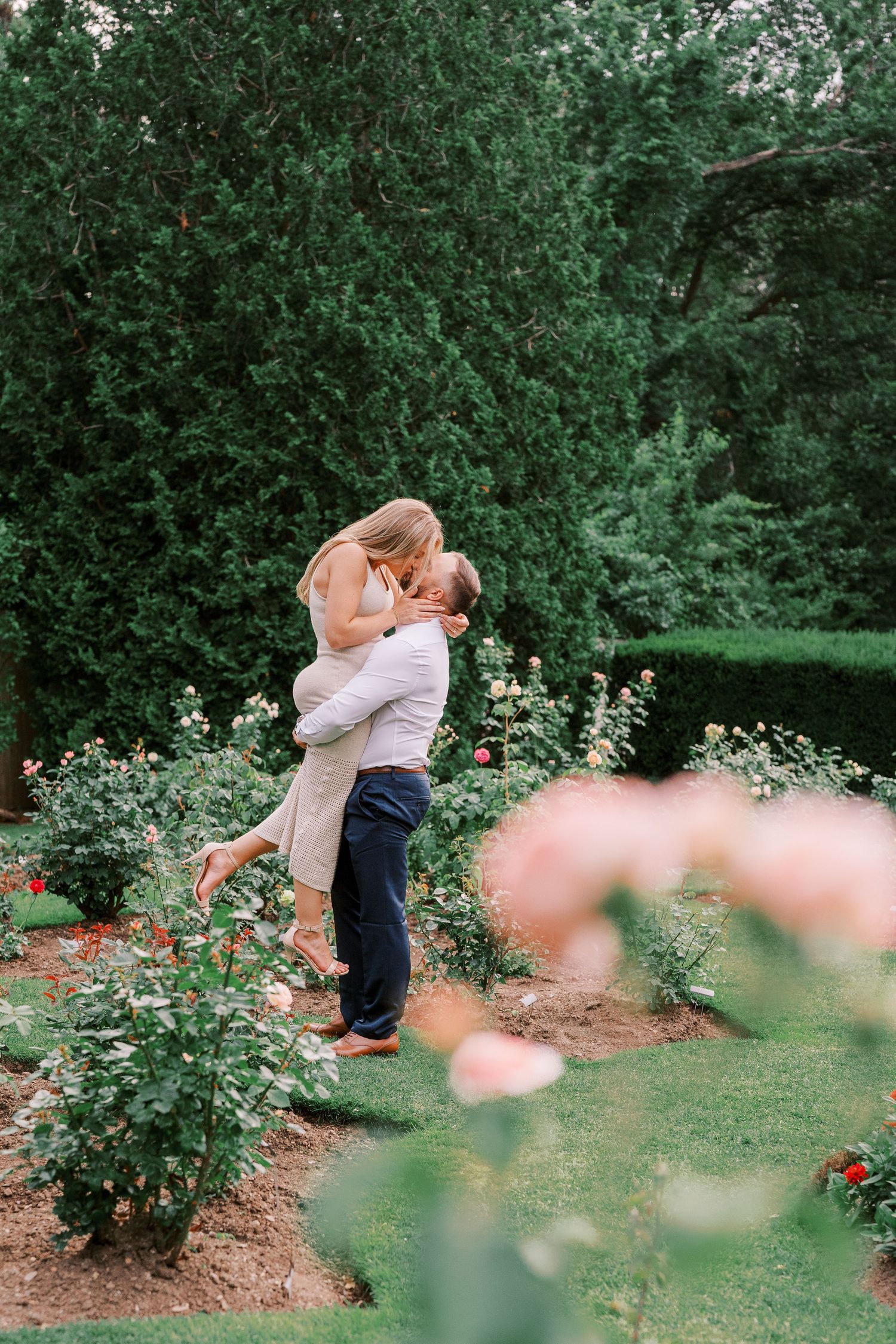 A romantic embrace between a couple in a lush garden surrounded by blooming roses and greenery.
