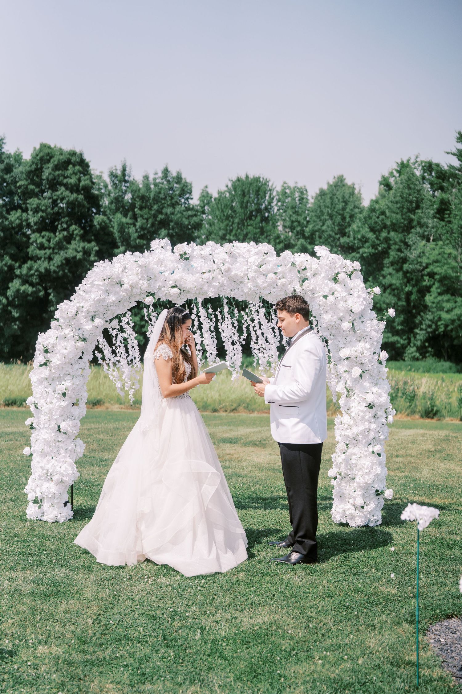 A beautiful outdoor wedding ceremony setup features an elegant white floral arch on a grassy field surrounded by trees.