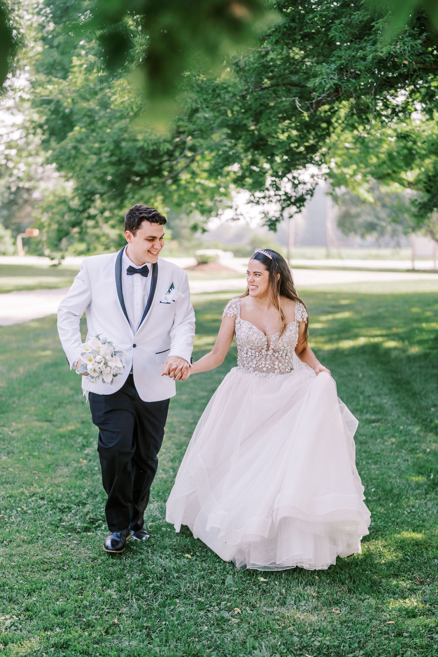 Newlyweds walk together on a lush green lawn, bride in flowing white gown and groom in white tuxedo jacket with bowtie.