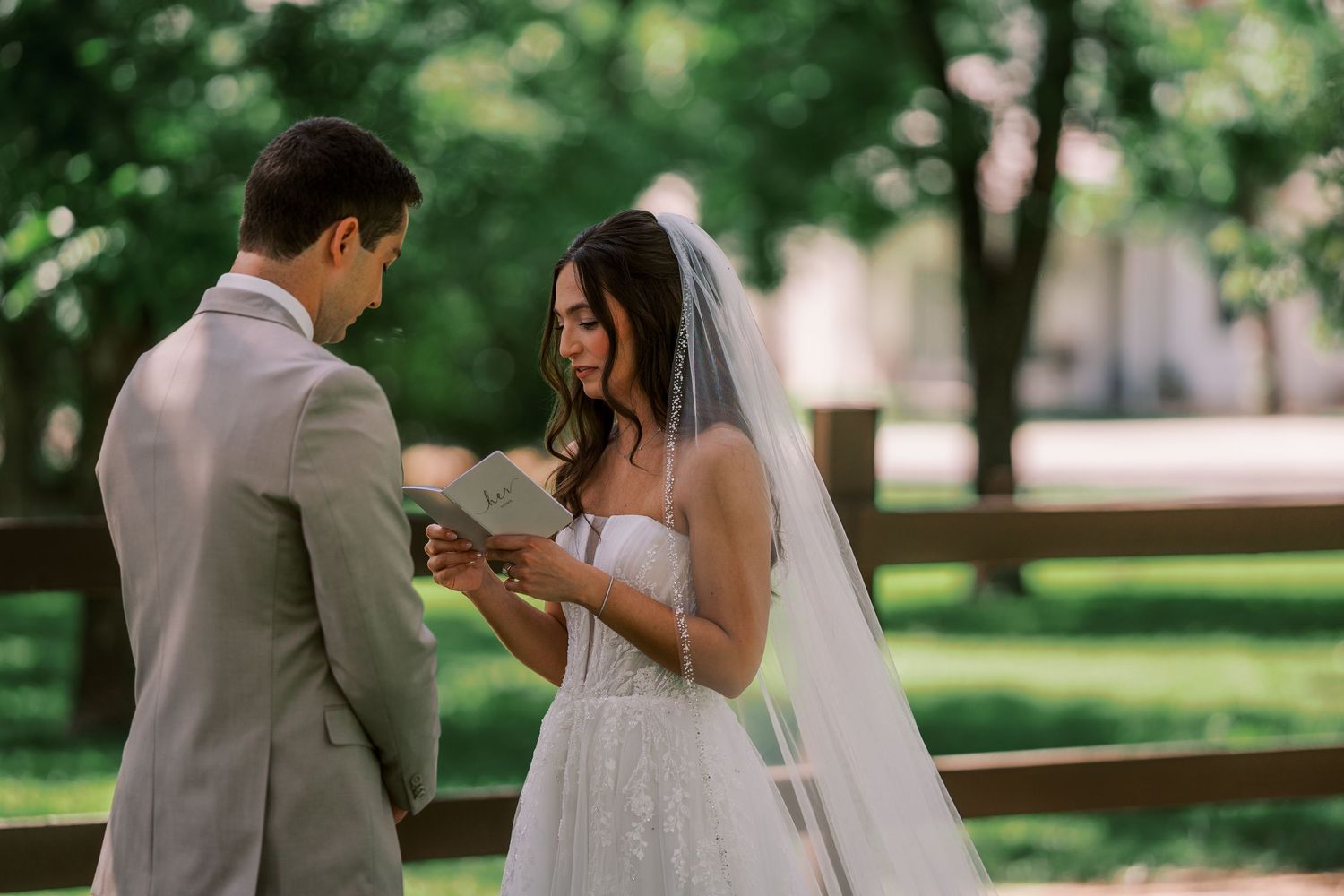 A couple exchanges rings during an outdoor wedding ceremony in a lush garden setting with soft natural lighting.