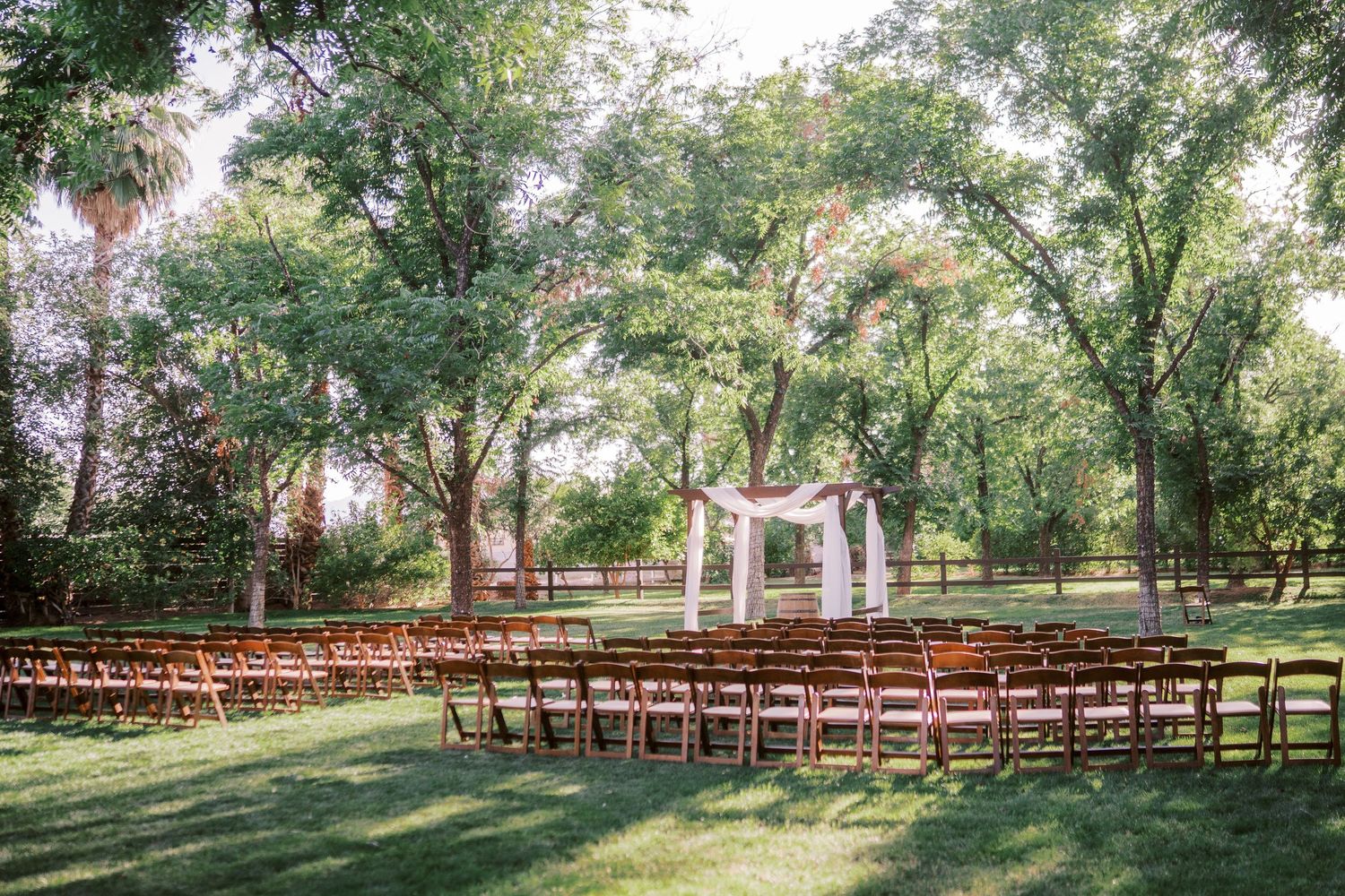 An elegant outdoor wedding ceremony setup with wooden chairs and a white arch surrounded by lush green trees.