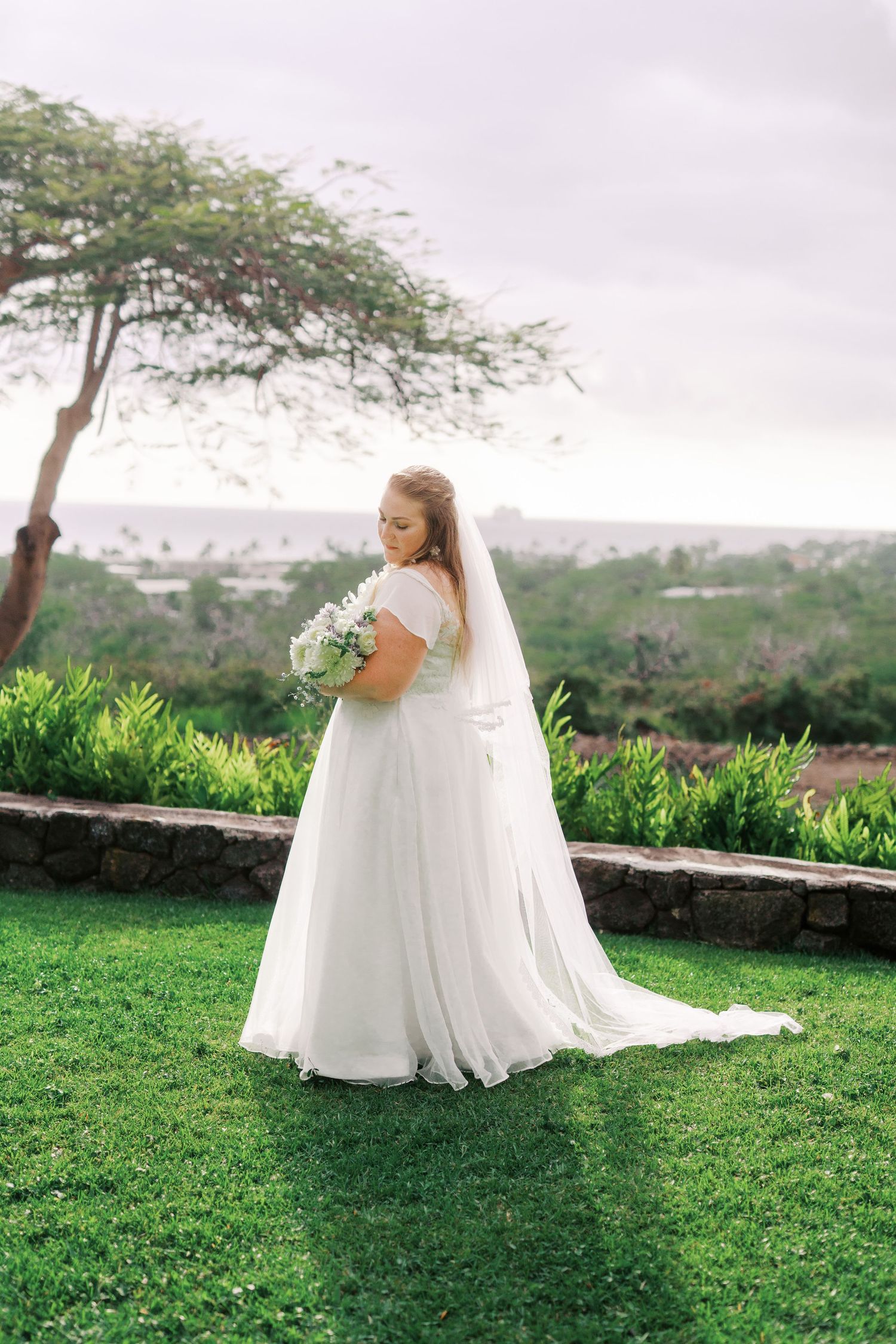 A bride in a flowing white dress poses on a scenic overlook with city views.
