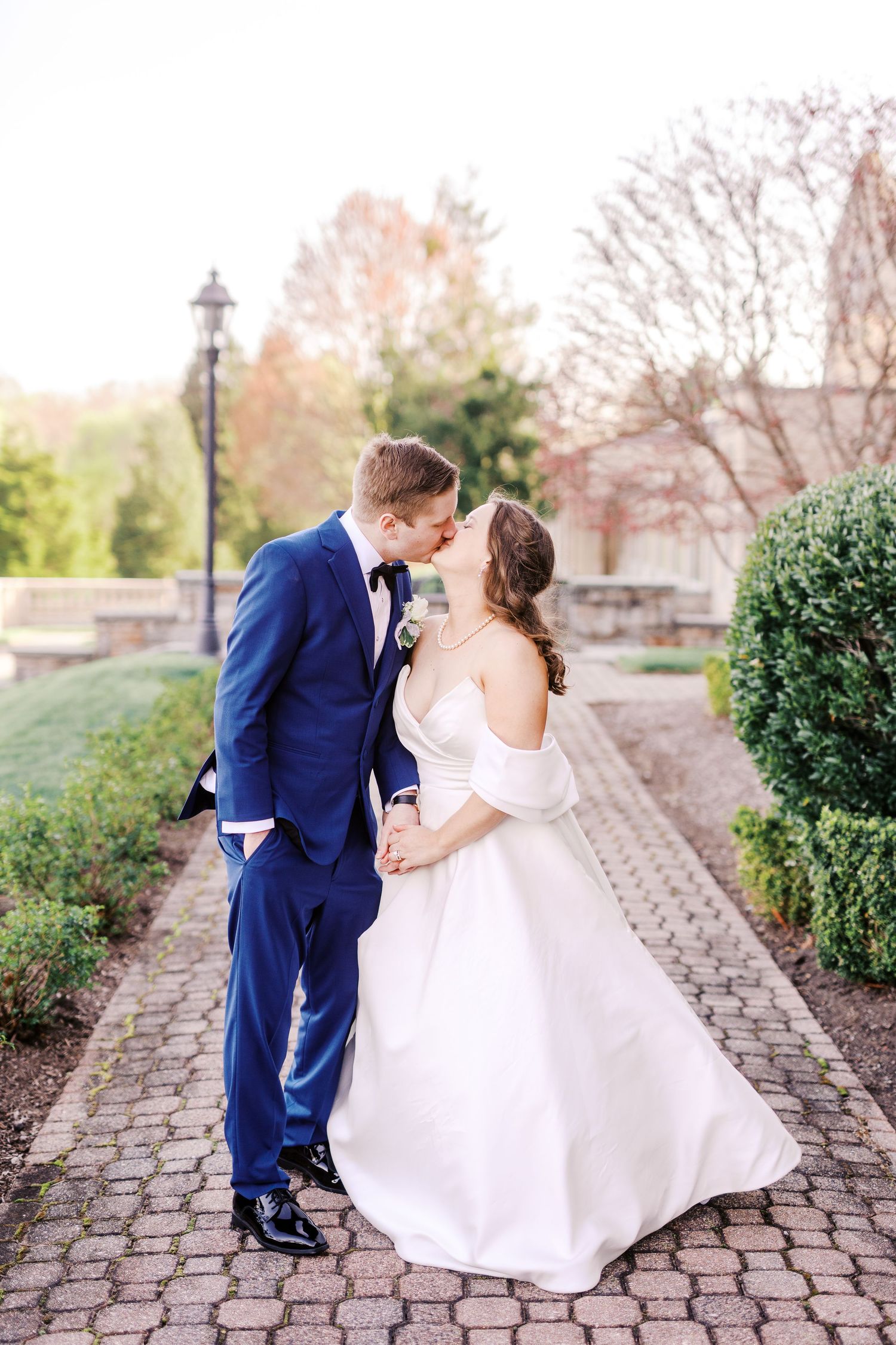 Newlyweds in formal attire share an intimate moment on a cobblestone path surrounded by garden landscaping.