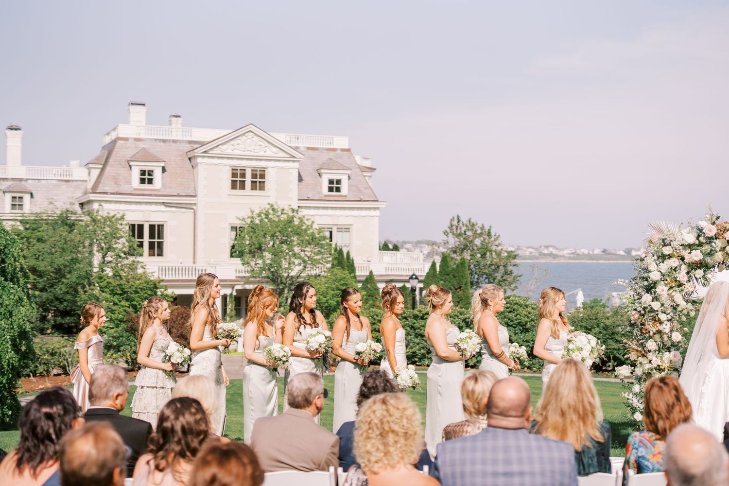A group of people gather outdoors for a celebratory event with a white building in the background at sunset.