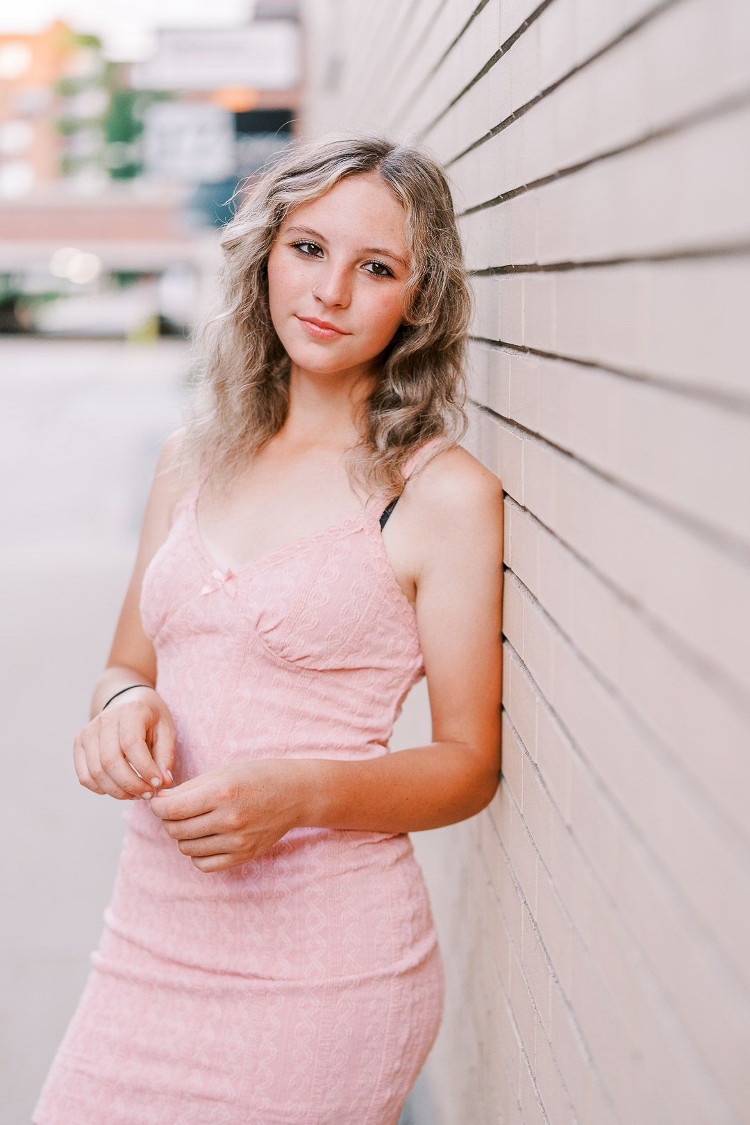 Young woman in pink dress poses against white wall outdoors in natural lighting.