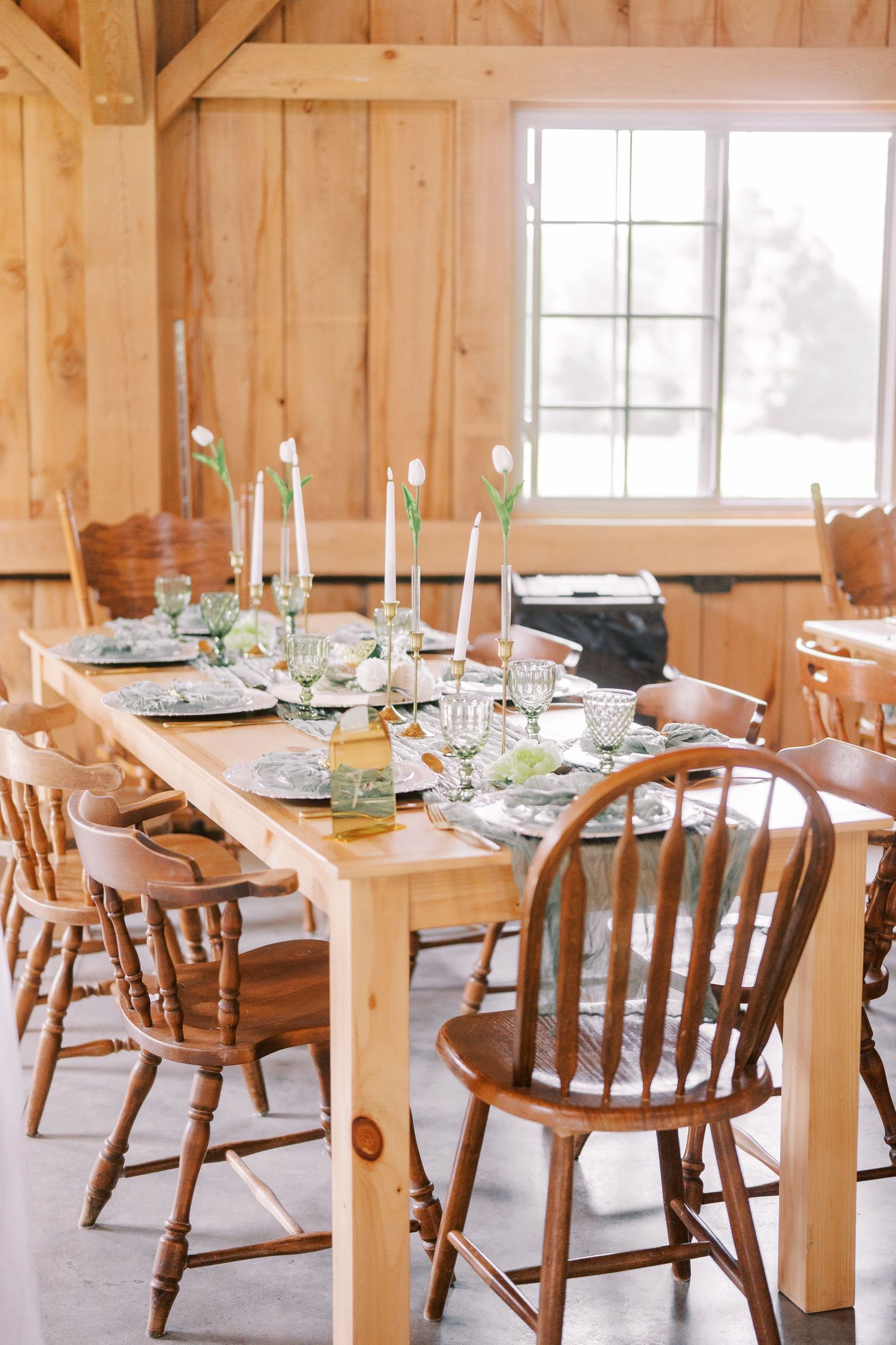 A rustic dining room interior with wooden tables and chairs set for a meal, featuring natural light from windows.