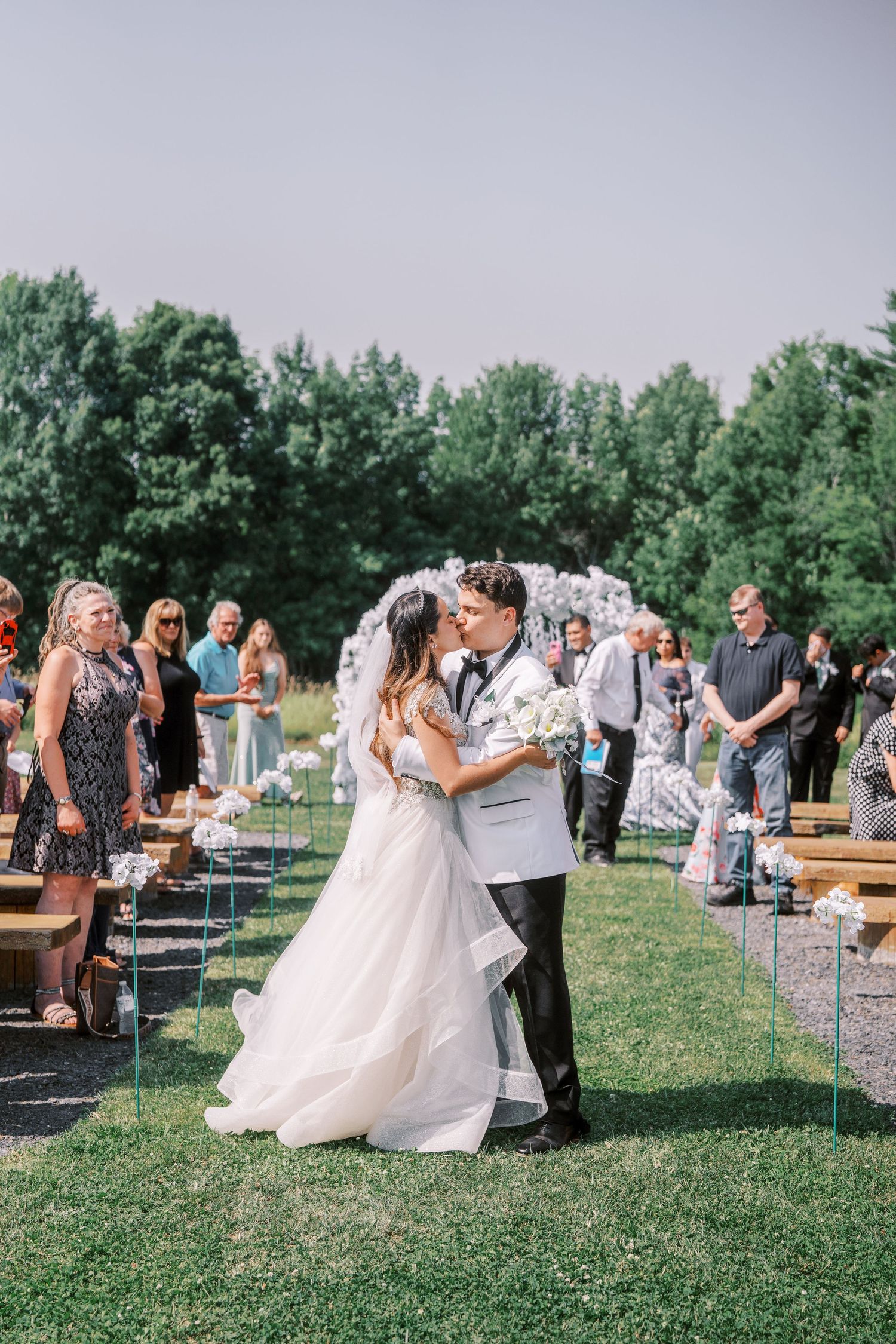 A romantic outdoor wedding ceremony with guests watching as a couple shares their first kiss as newlyweds on a grassy lawn.