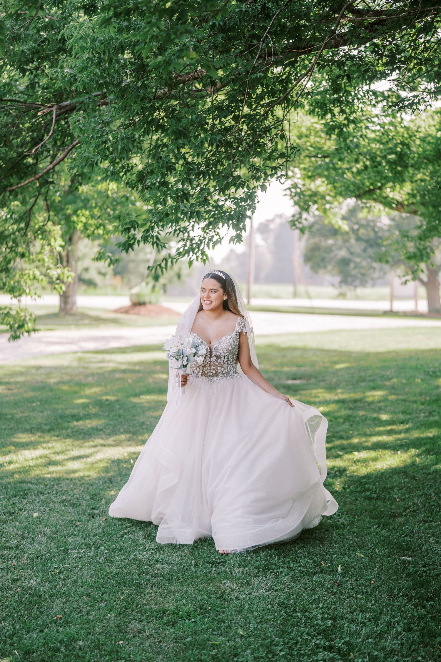 A flowing white wedding dress billows gracefully across green grass in a sunlit outdoor garden setting.