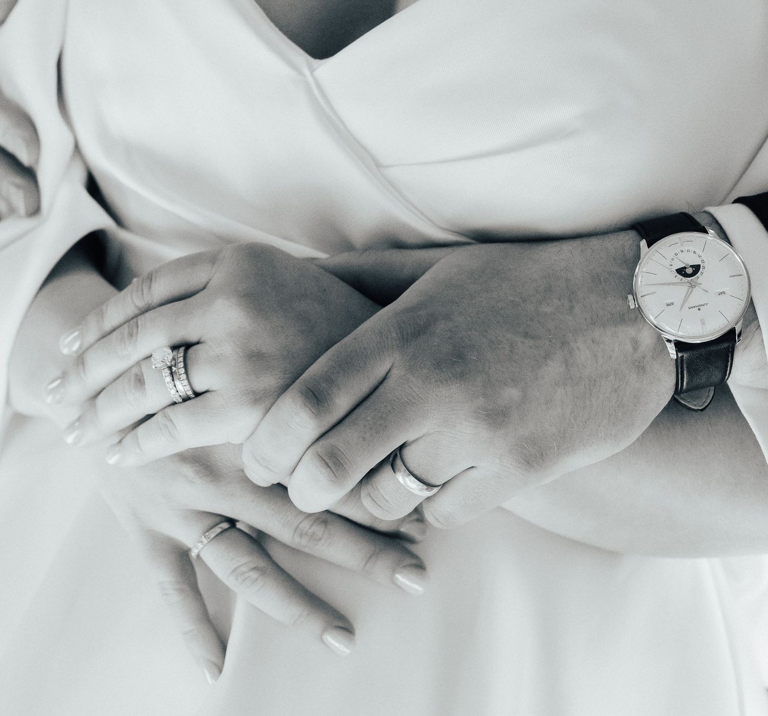 Black and white close-up of hands wearing rings and a wrist watch against soft white fabric.