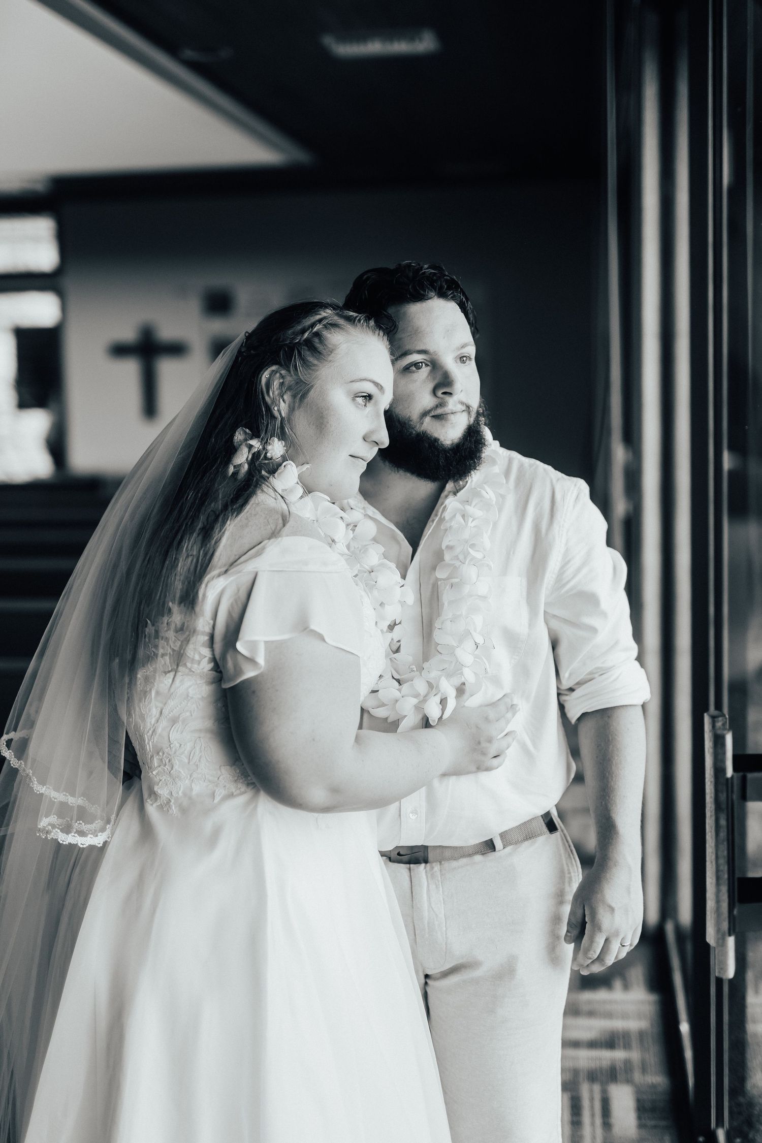 Intimate black and white wedding portrait of couple embracing near a window with a church cross in the background.