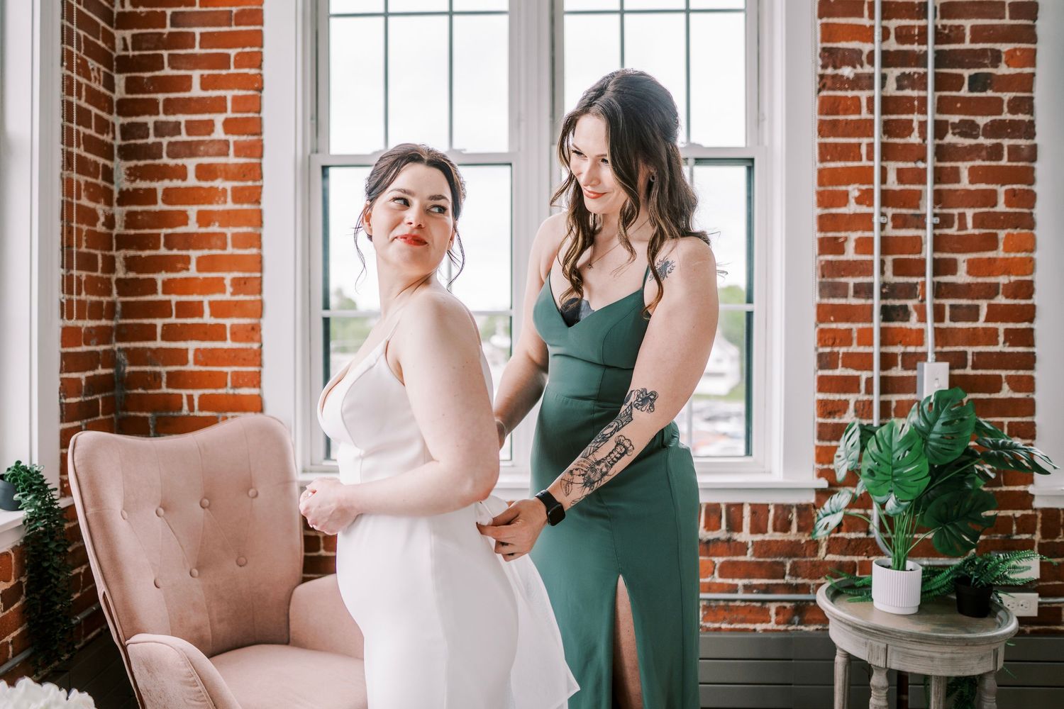 Two people in formal dresses, one in white and one in emerald green, stand together in a rustic brick-walled room.