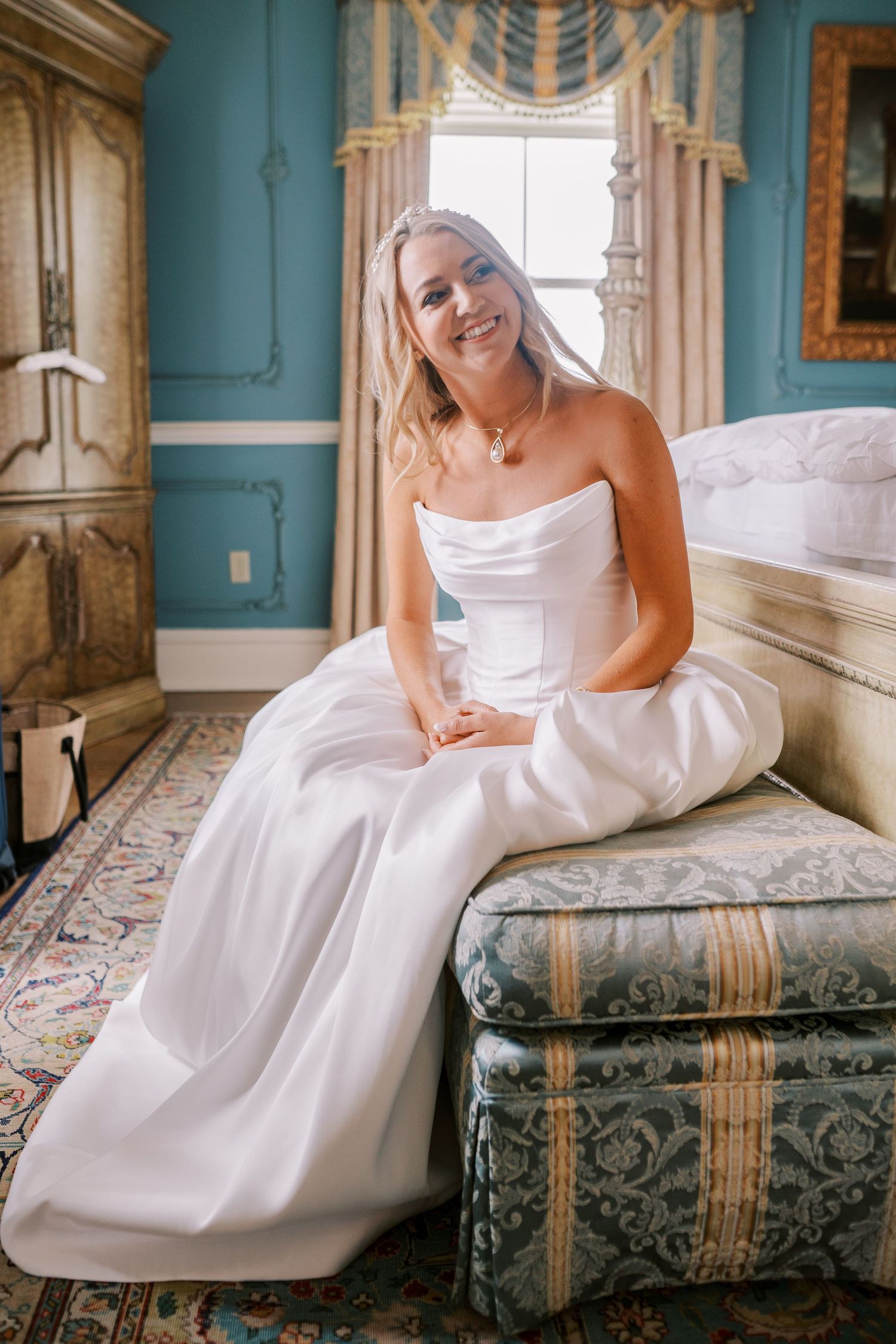 A white wedding dress drapes elegantly on an antique settee in a turquoise-walled room with ornate window treatments.
