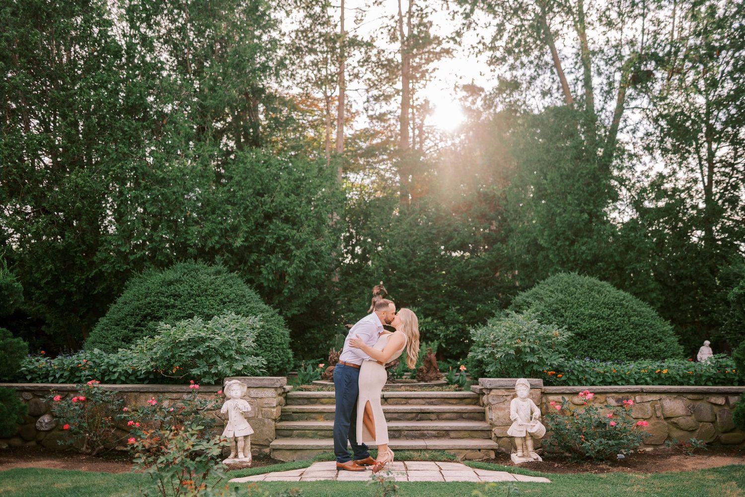 A couple embraces on garden steps at sunset with perfectly trimmed hedges and trees in the background.