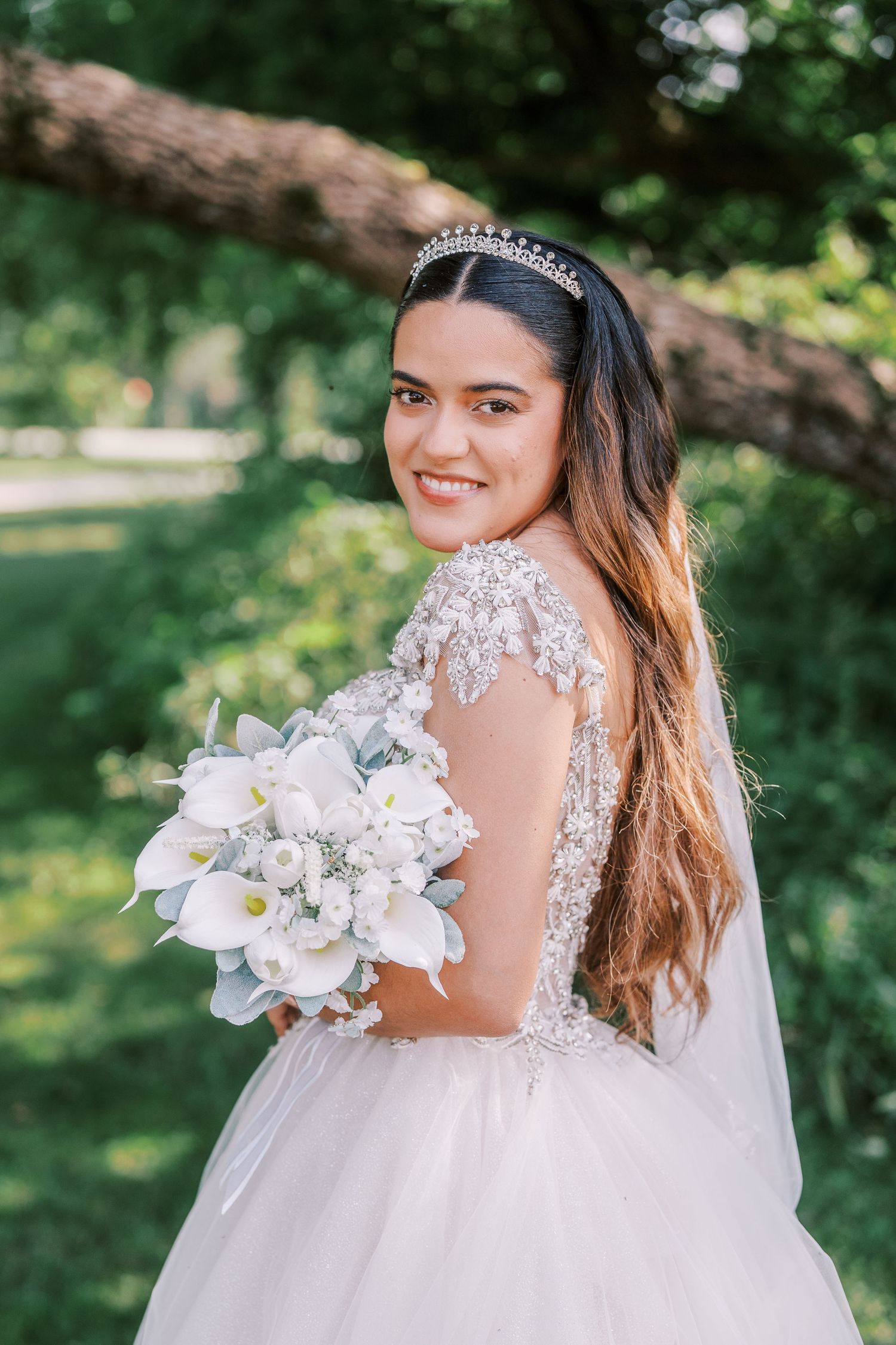 Beautiful bride in lace gown with crystal tiara holds white calla lily bouquet in outdoor garden setting.