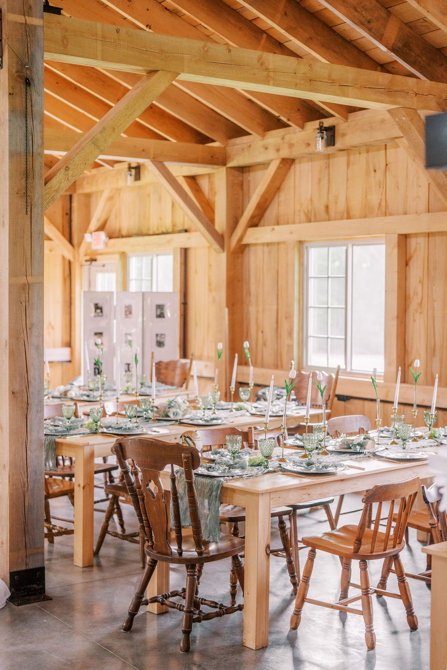 Rustic barn dining room with wooden tables and chairs set for a wedding reception under exposed timber beams and natural light.