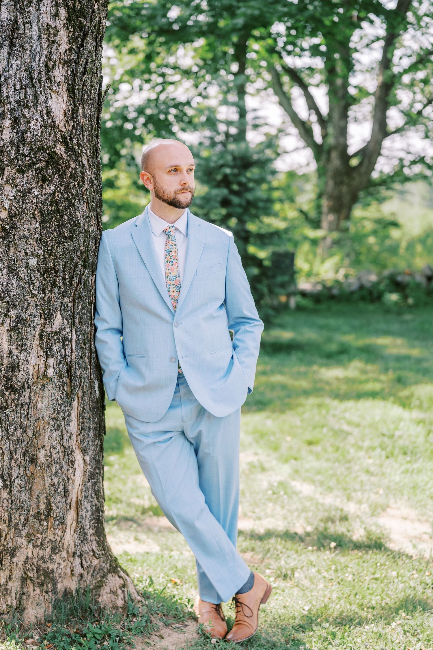 A man in a light blue suit and floral tie leans casually against a tree in a sunny outdoor setting.