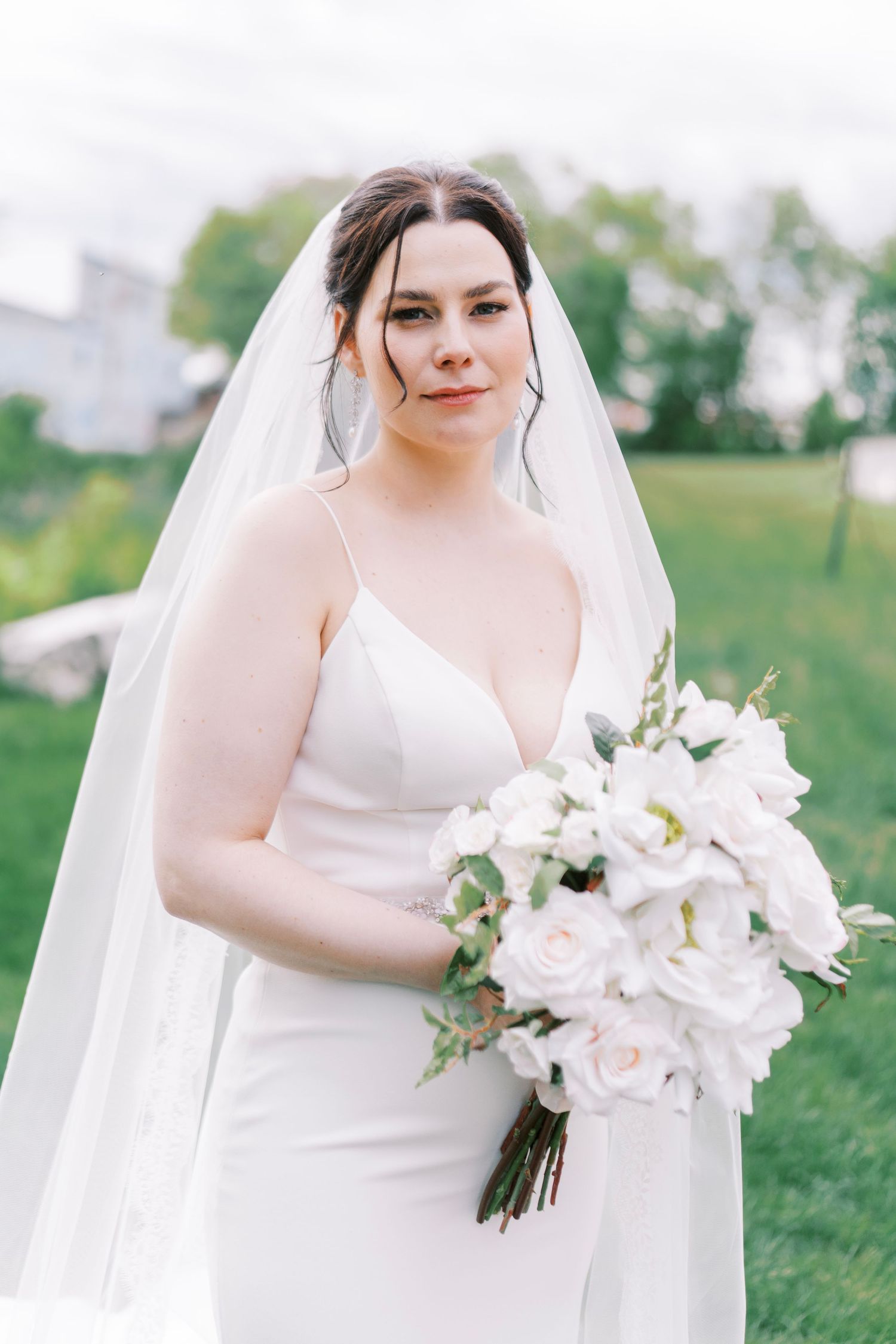 A bride in a sleek white wedding dress holds a stunning bouquet of white orchids and roses in an outdoor garden setting.