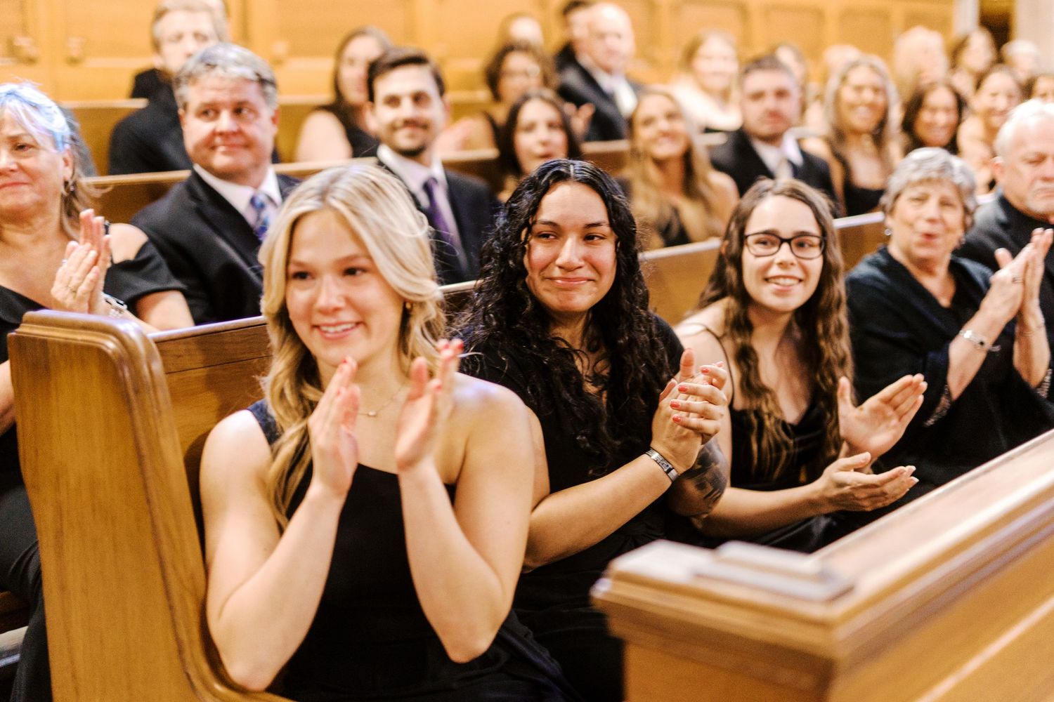 Wedding guests seated in wooden church pews smile and clap during a joyous ceremony celebration.