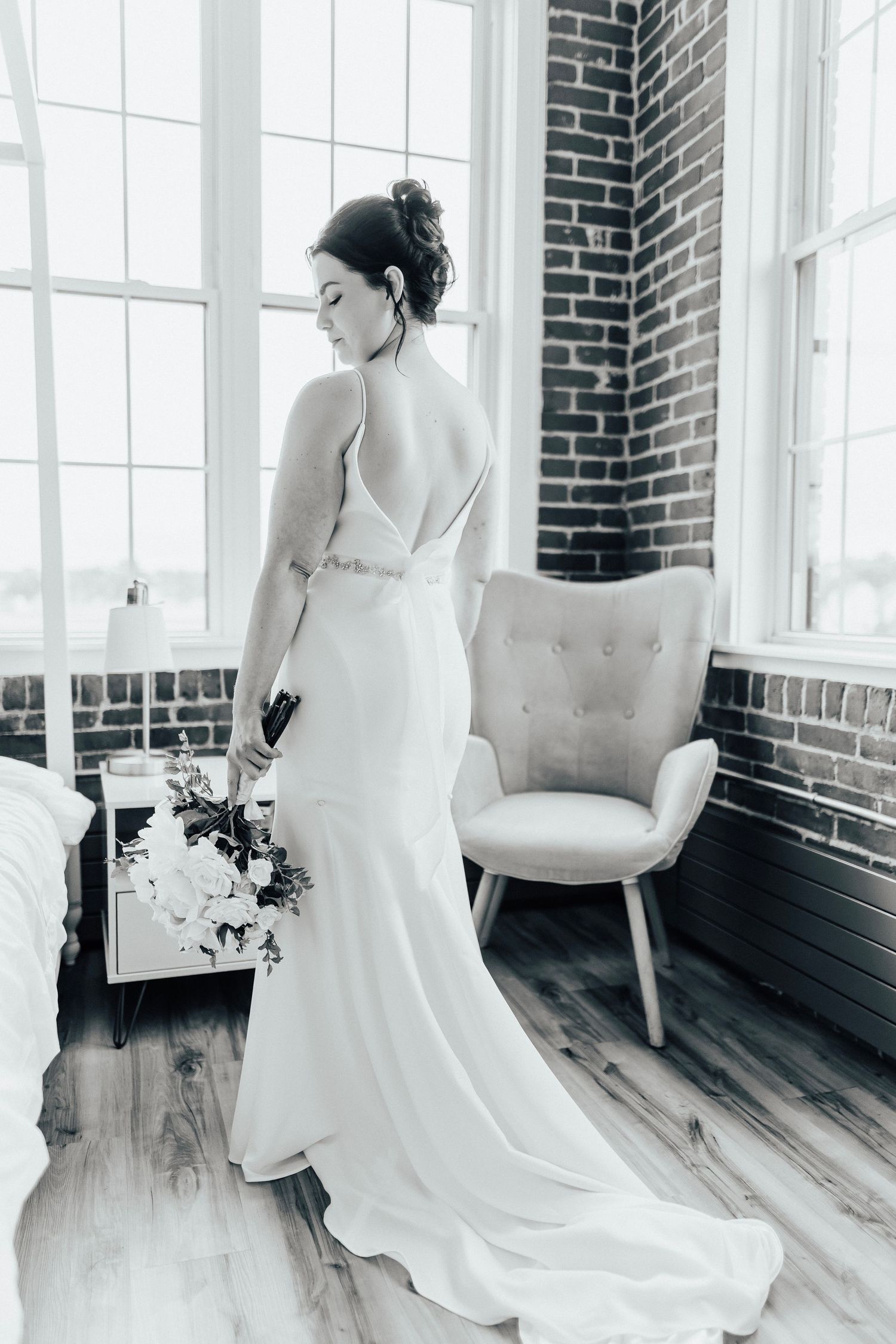 Bride in elegant white fitted dress poses near brick wall and window.