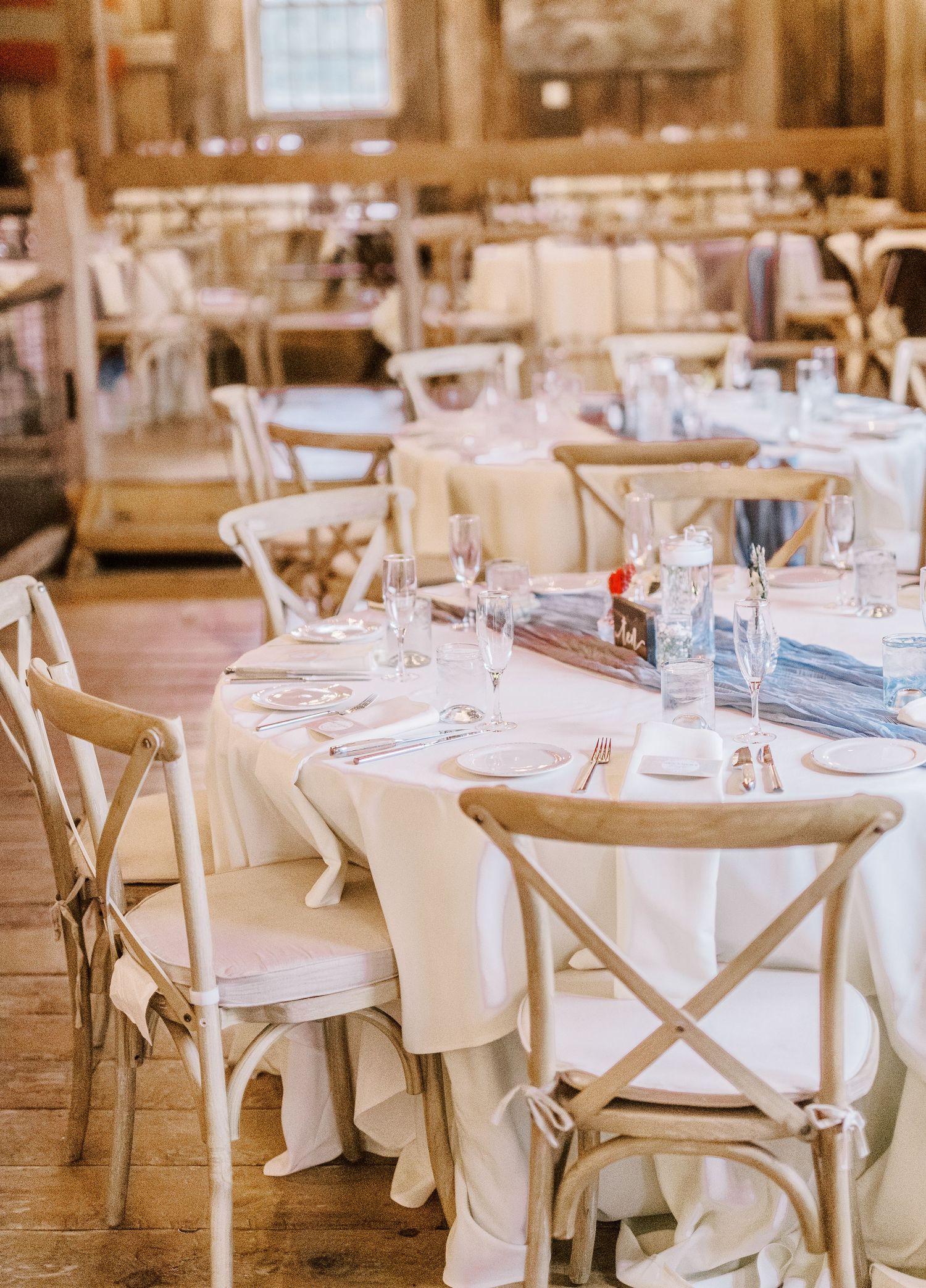 Elegant wedding reception venue with white-clothed tables and cross-back chairs in a rustic indoor setting.