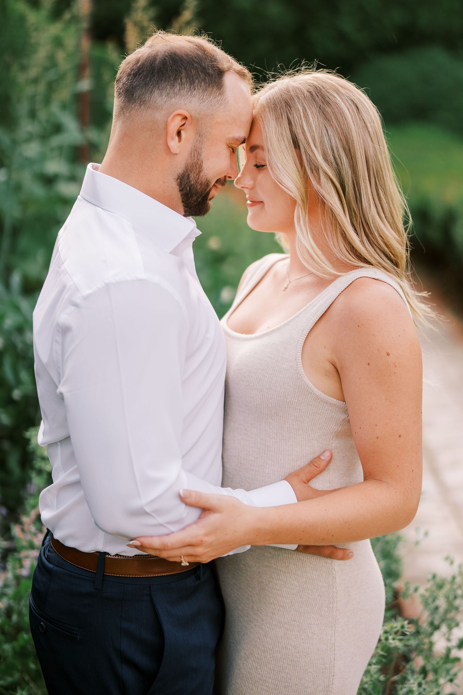 A romantic couple embraces lovingly in an outdoor garden setting with lush greenery in the background.