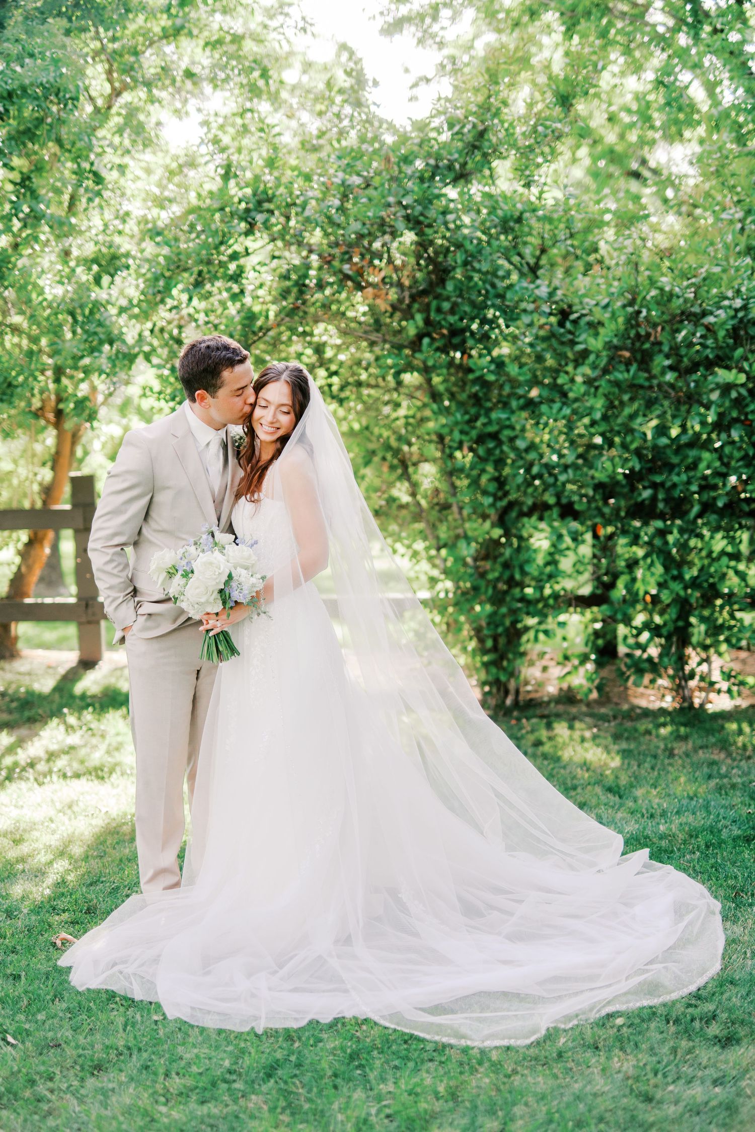 A wedding couple embraces in a sunlit garden setting with a flowing white dress and veil.