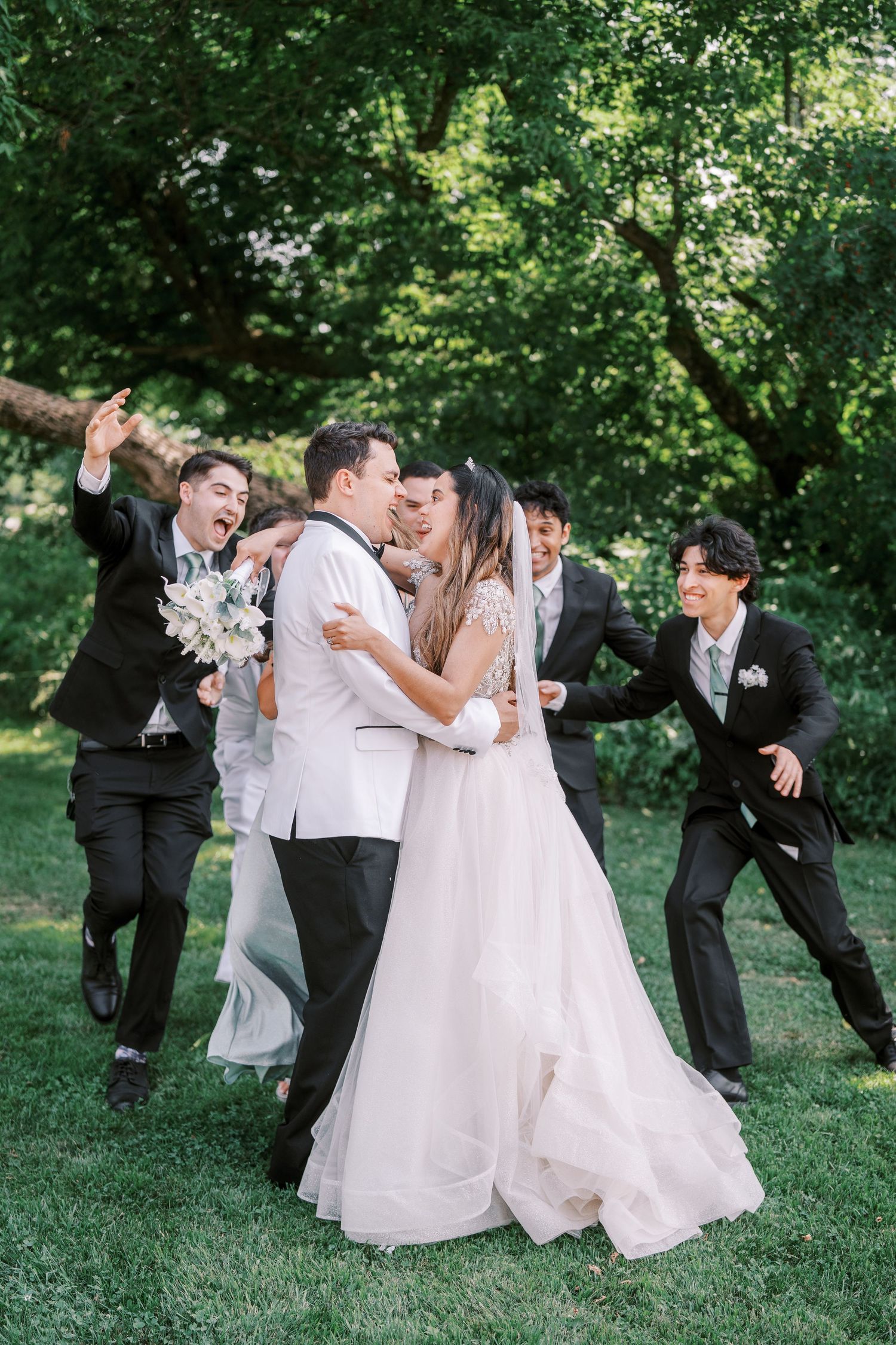 Newlywed couple shares romantic kiss in garden setting while wedding party celebrates joyfully behind them.