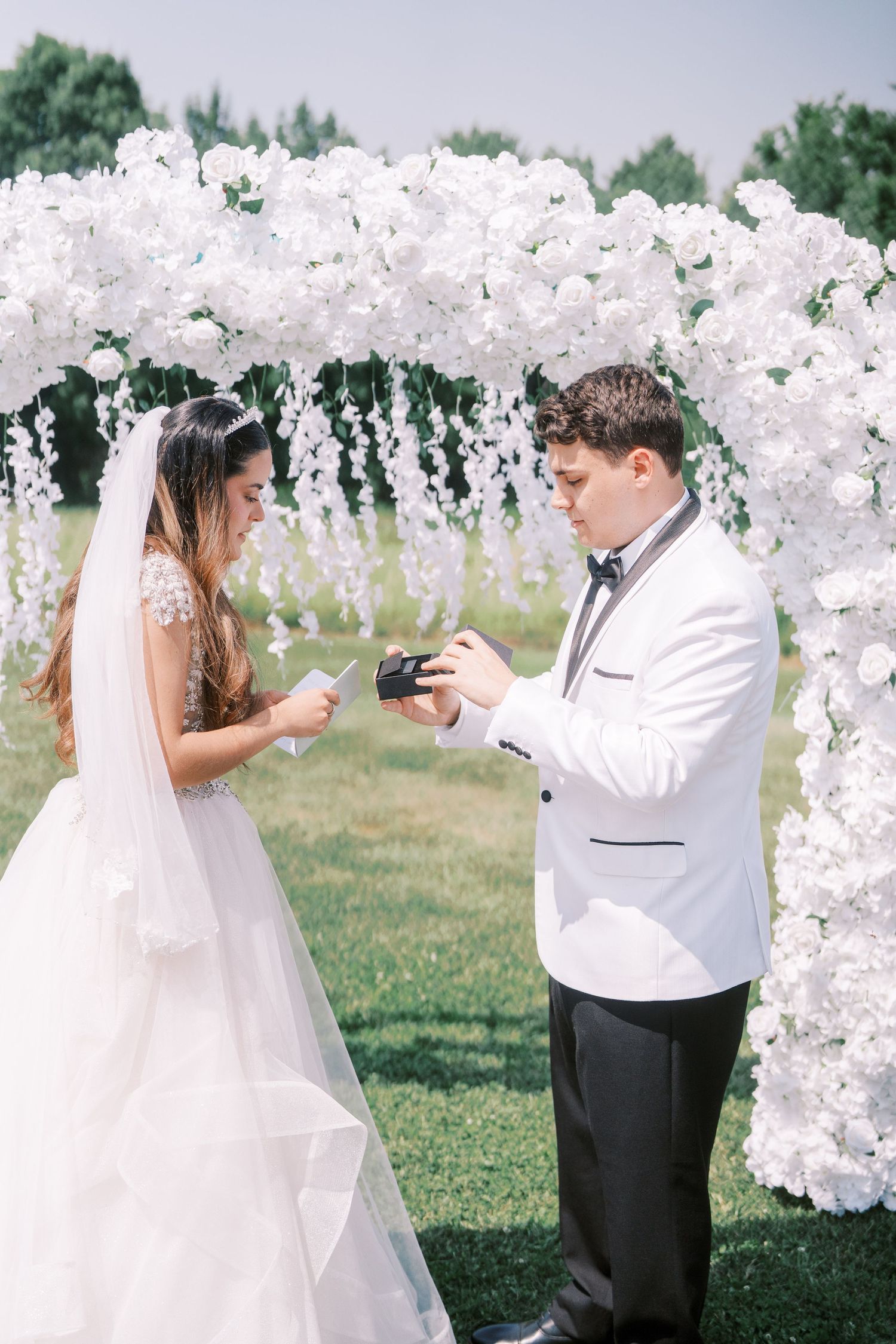 A couple exchanges wedding vows under a white floral arch adorned with cascading blossoms in an outdoor garden setting.