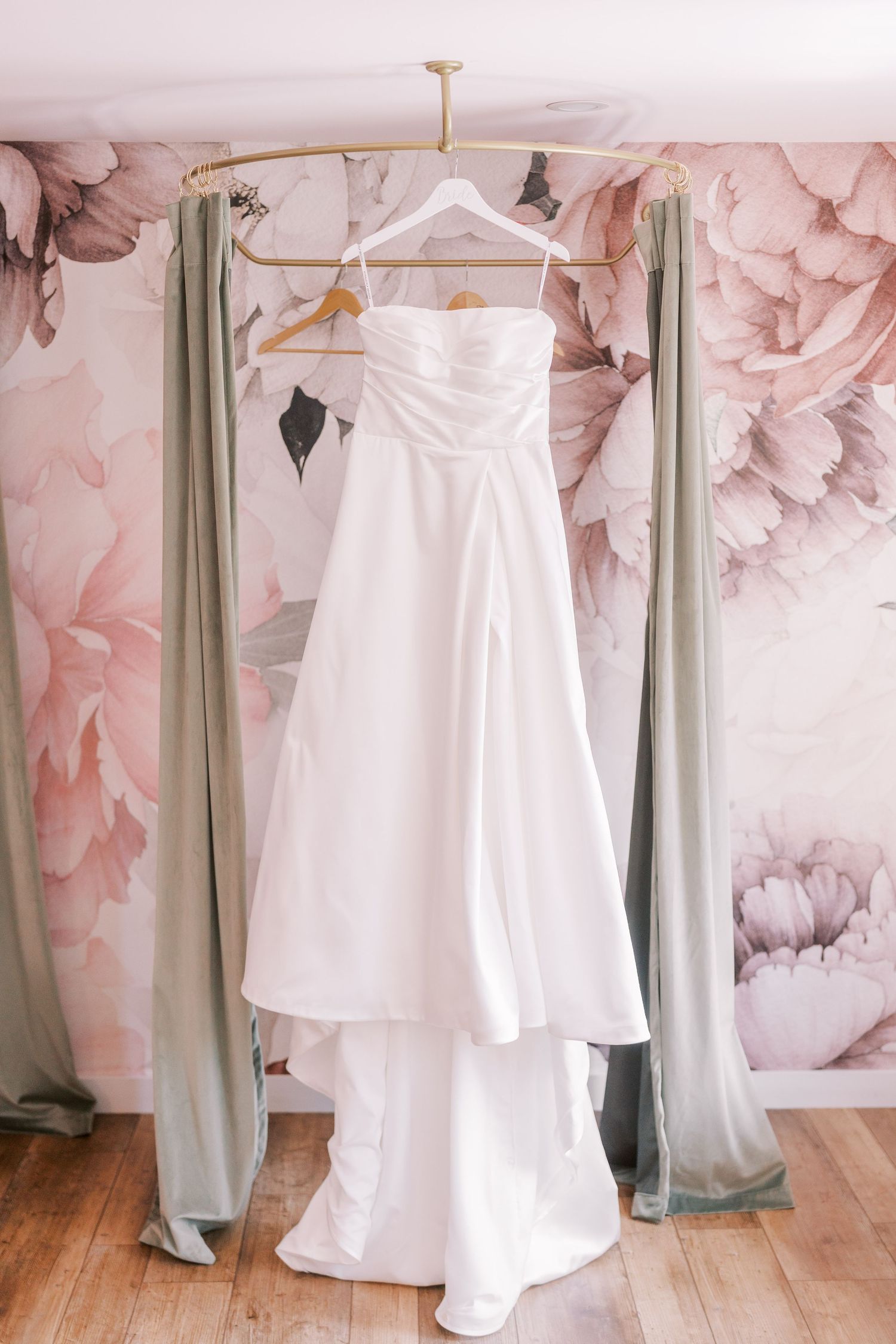 Elegant white wedding dress hanging against a pink floral wallpaper with sage green curtains.