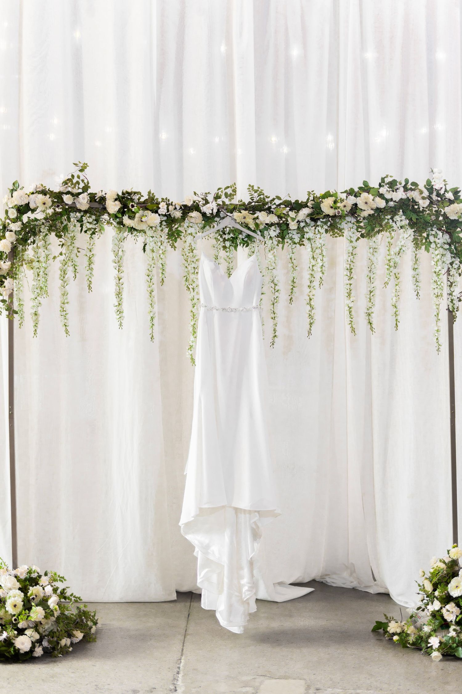 Elegant white wedding dress hanging beneath a floral arch with cascading greenery and white flowers.