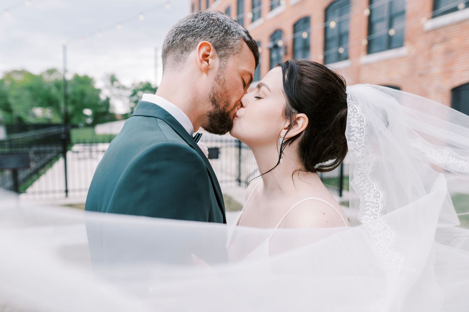 Romantic wedding kiss captured through delicate veil with brick building in background.