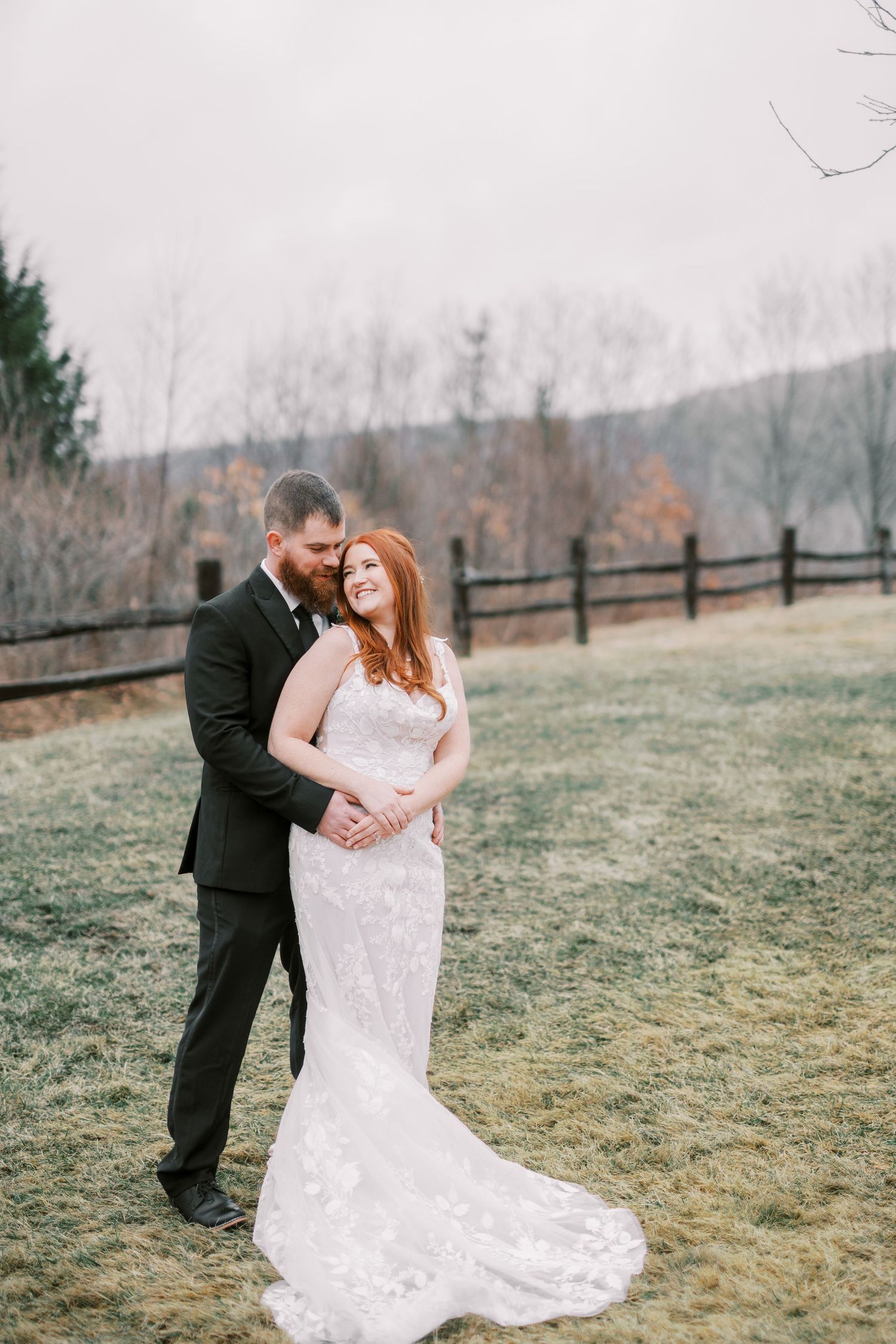A wedding couple embraces on a rustic property with a wooden fence and misty mountain landscape in the background.