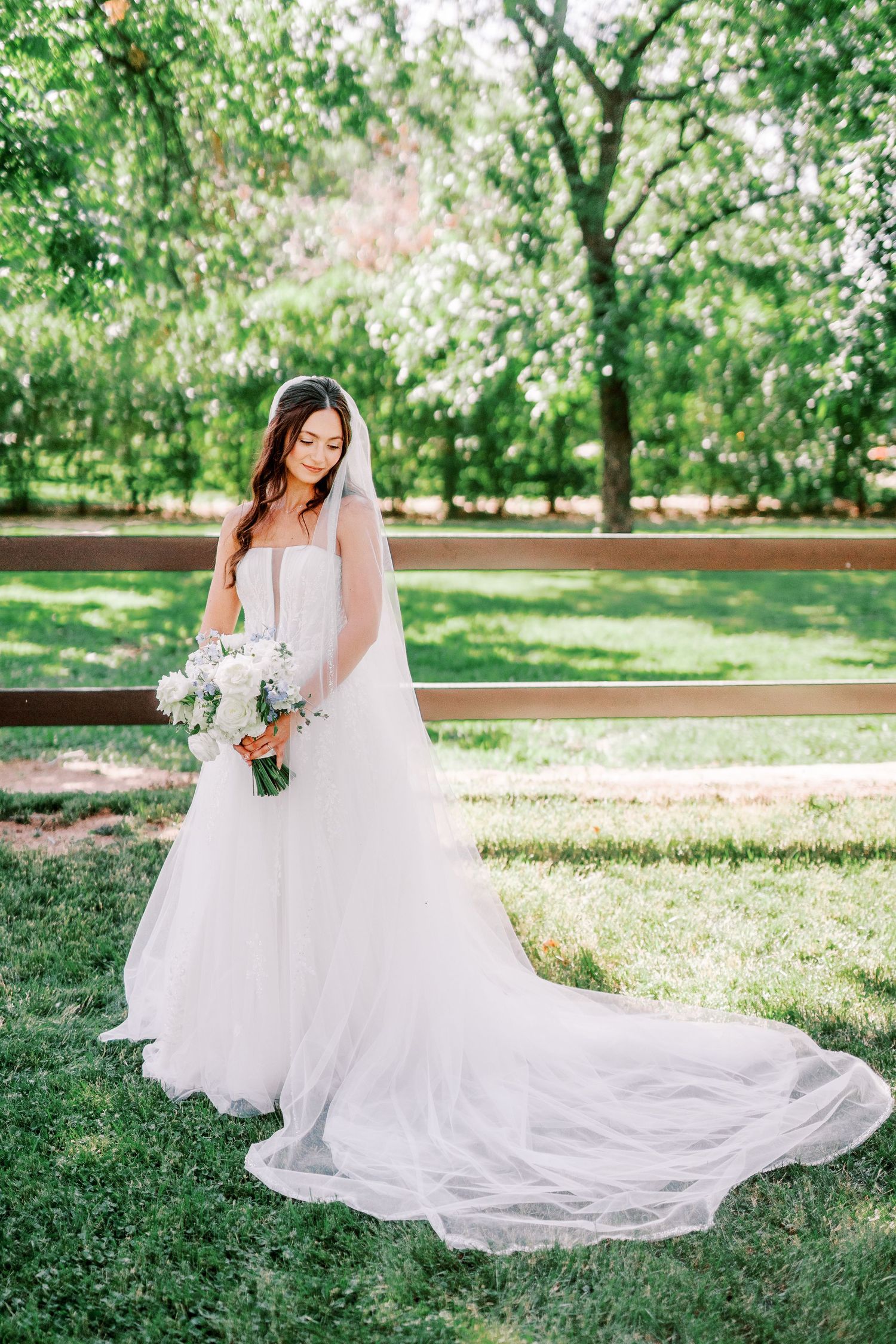 A bride in a flowing white wedding gown with long train holds a bouquet while standing in a sunlit garden setting.