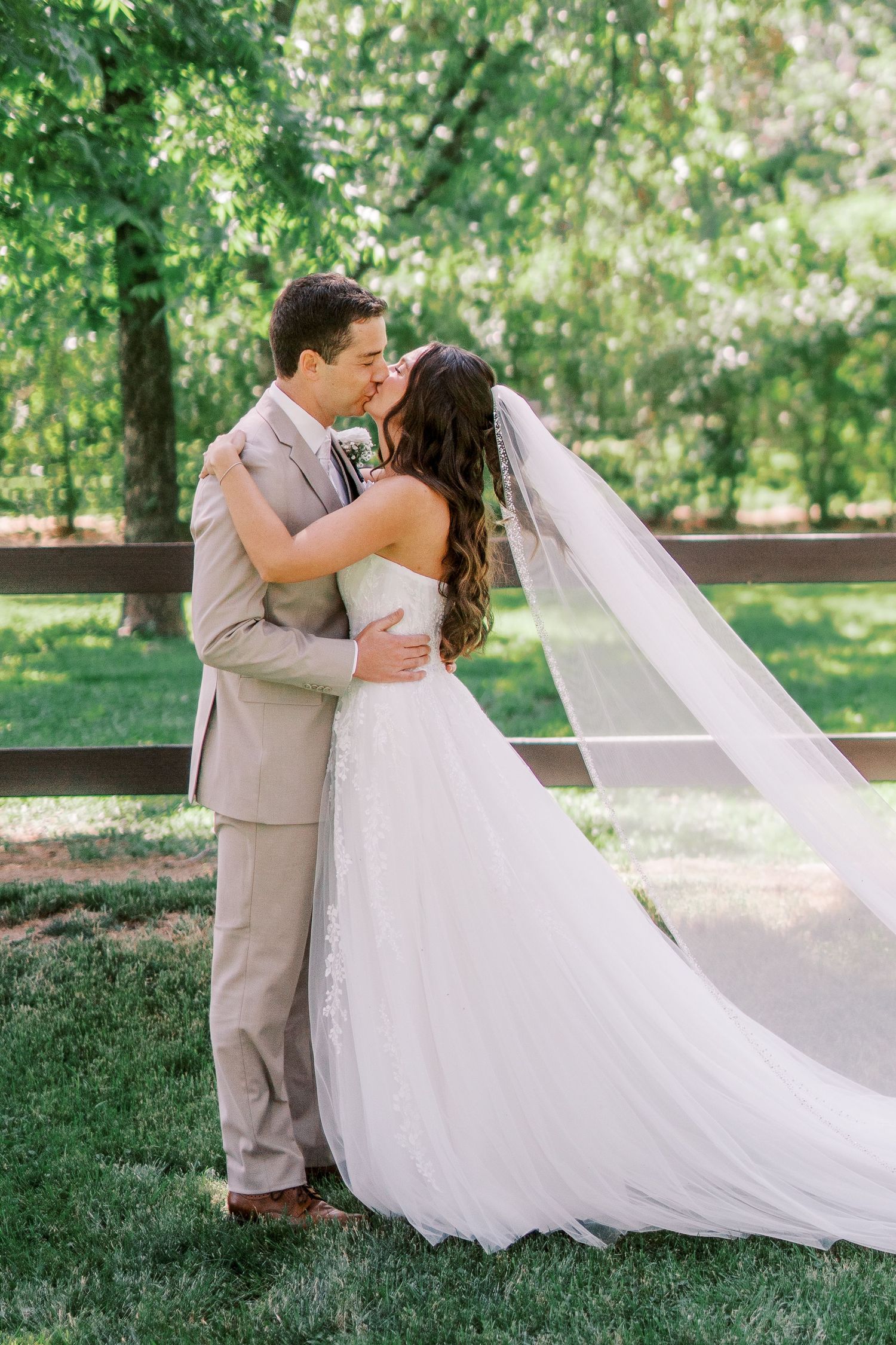 A couple shares intimate moments on the grass while the bride's long white veil flows elegantly in the outdoor garden setting.