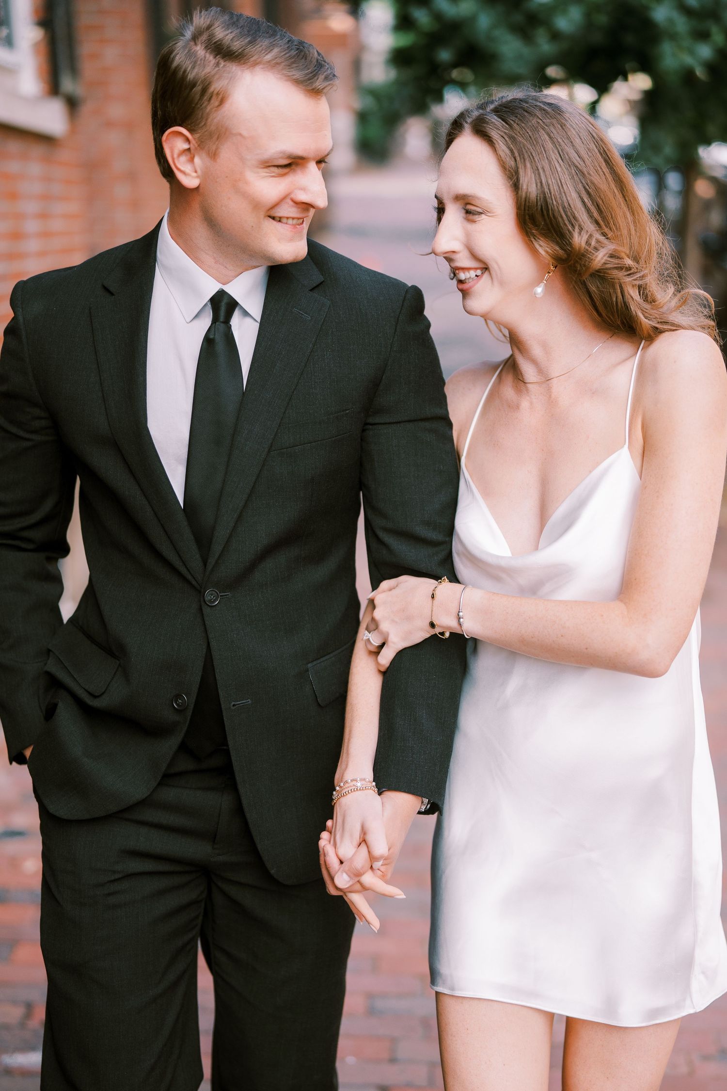 A couple in wedding attire laughs together while walking beside a brick wall during their wedding photoshoot.