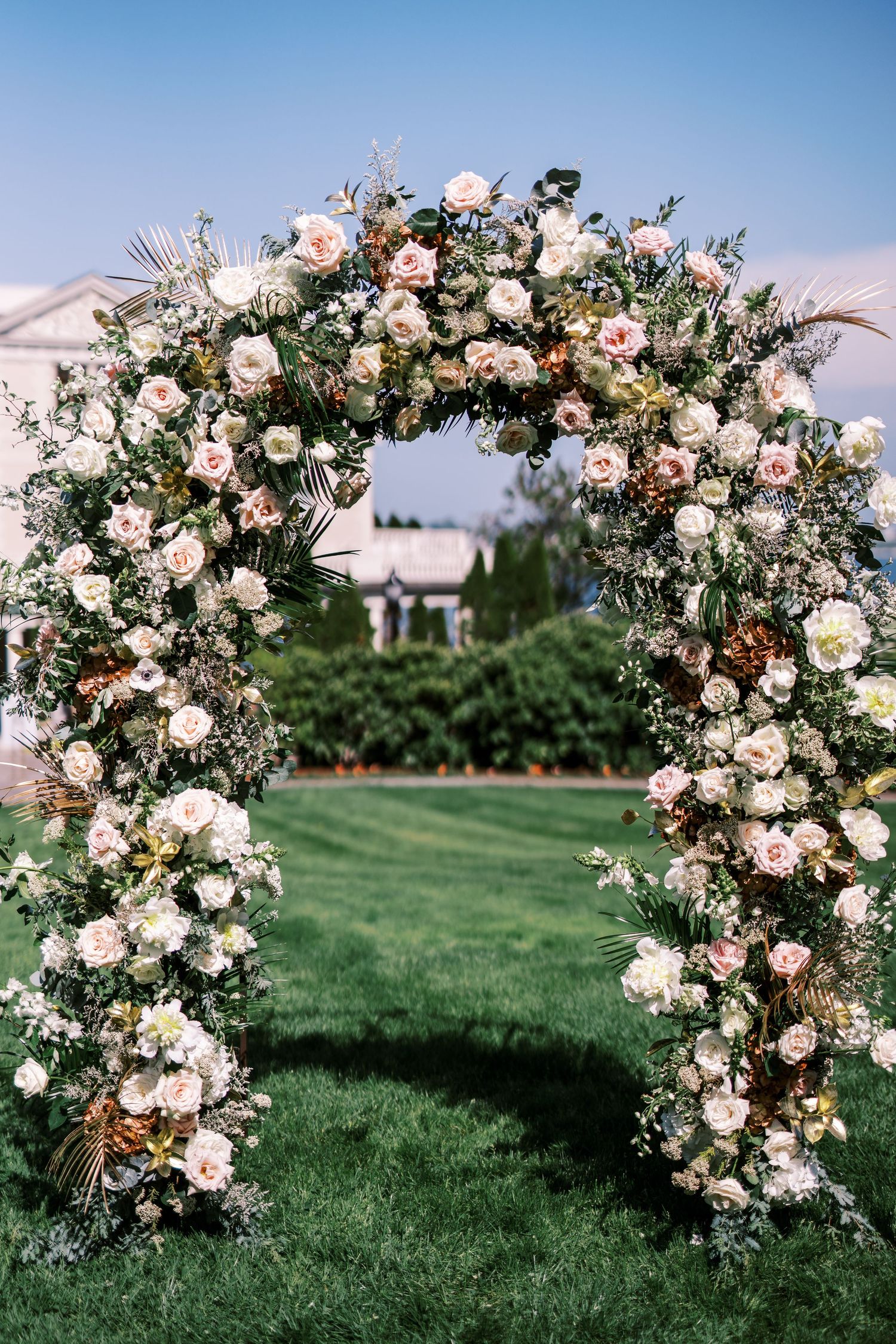 A romantic floral wedding arch covered in white roses and greenery stands elegantly on a lush green lawn against a blue sky.