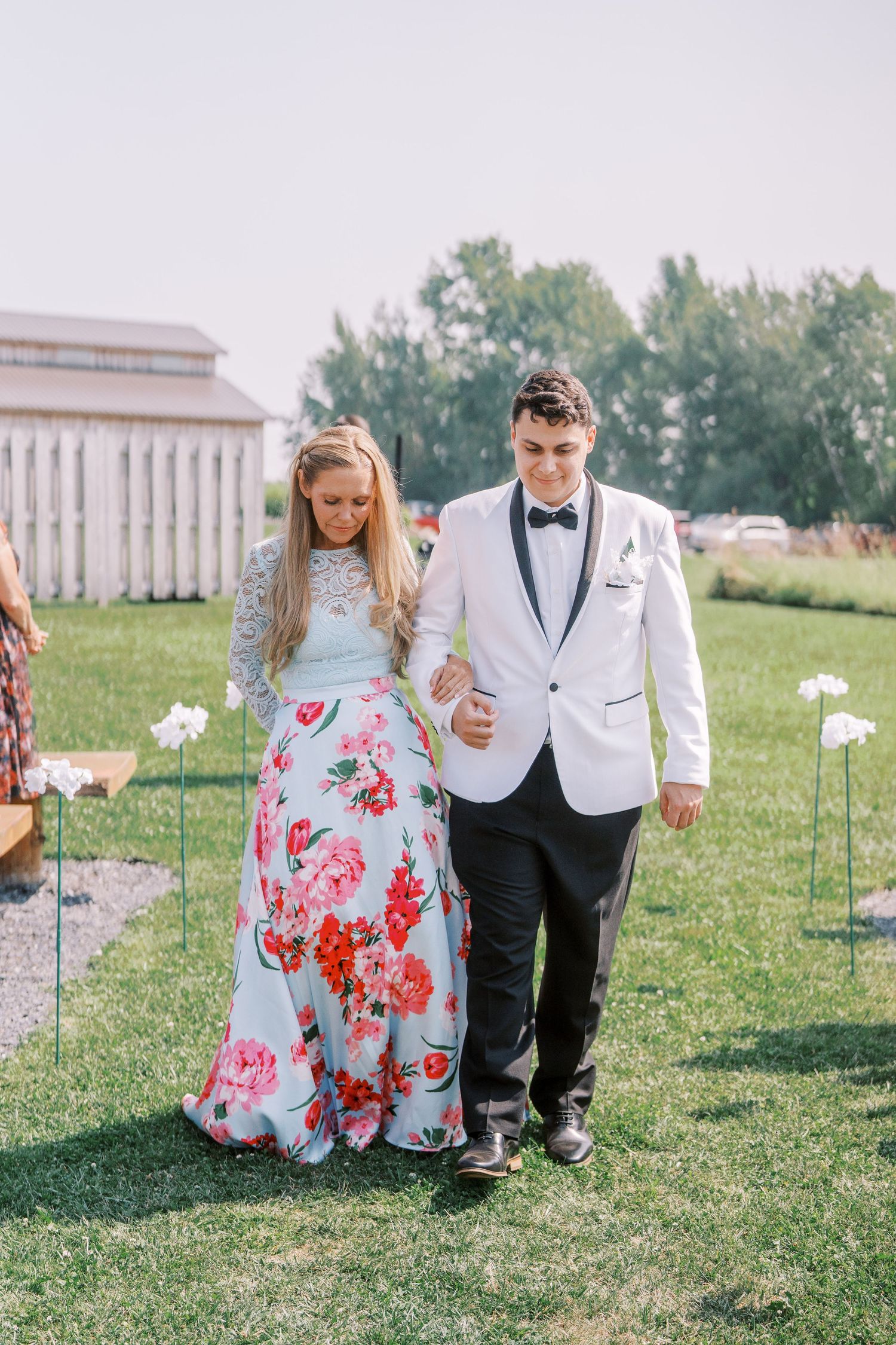 A couple in wedding attire stands together on a grassy lawn decorated with white floral accents.